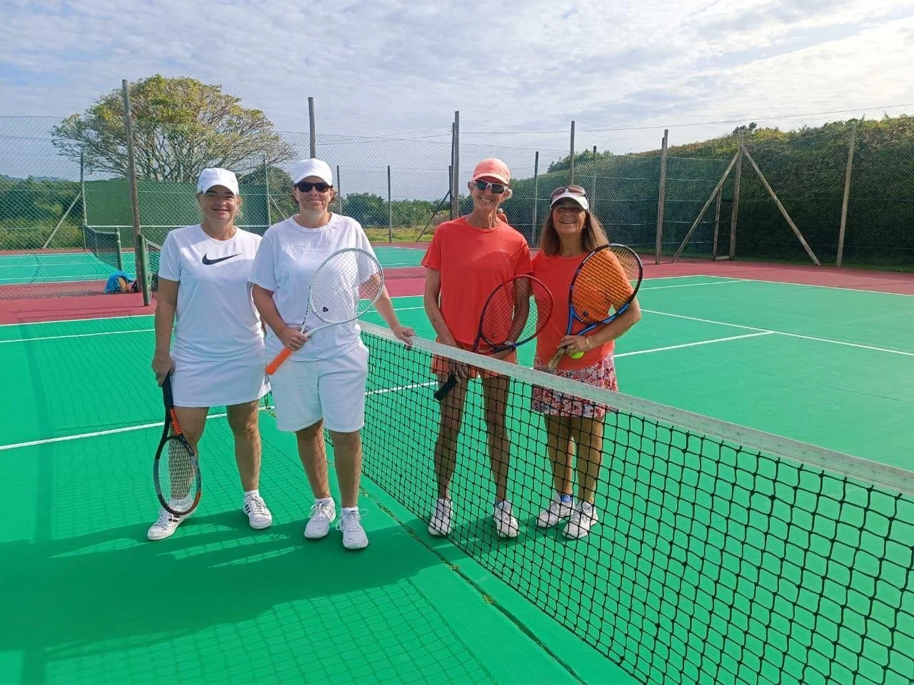 Four women standing on a tennis court behind a net, holding tennis rackets and smiling, with a green and red court and a blue sky with clouds in the background.