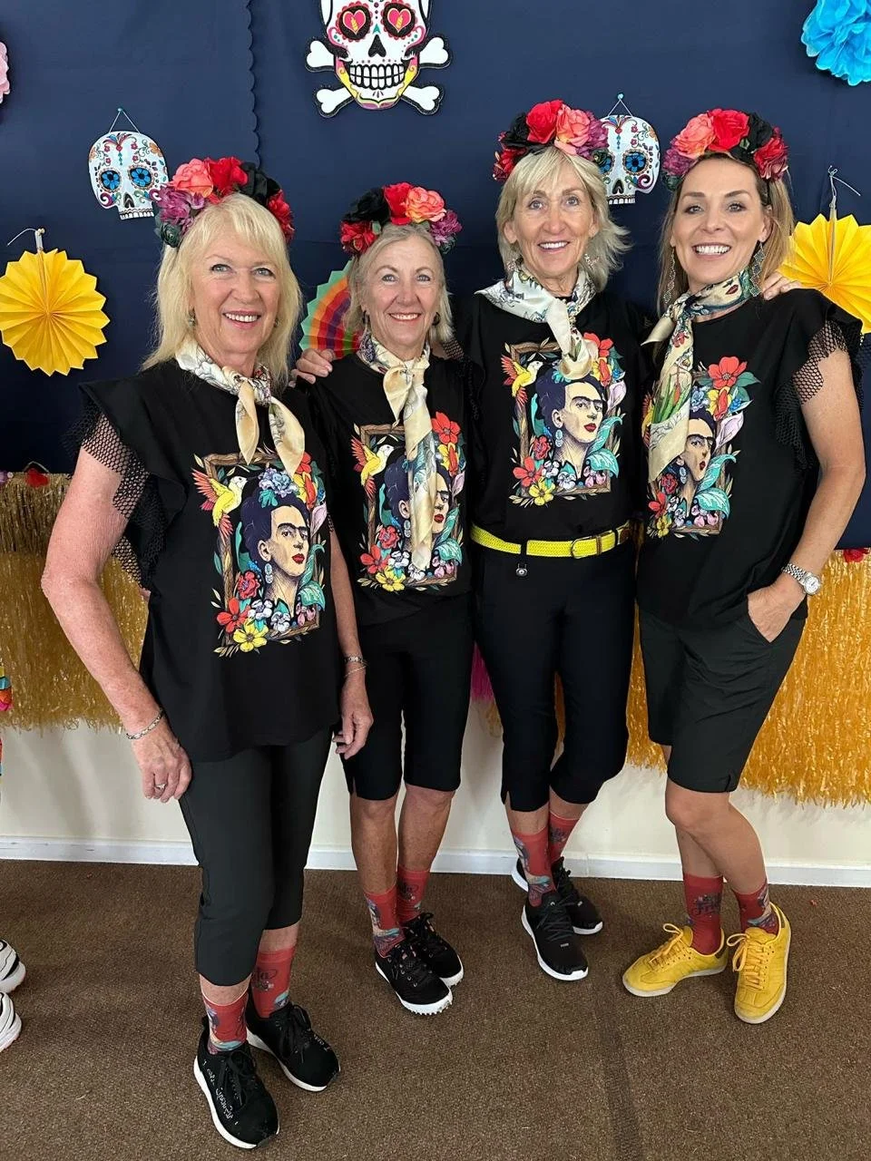 Four women dressed in black t-shirts with colorful Frida Kahlo designs, wearing floral headbands and bandanas around their necks, standing together in front of a Día de los Muertos themed decoration backdrop.