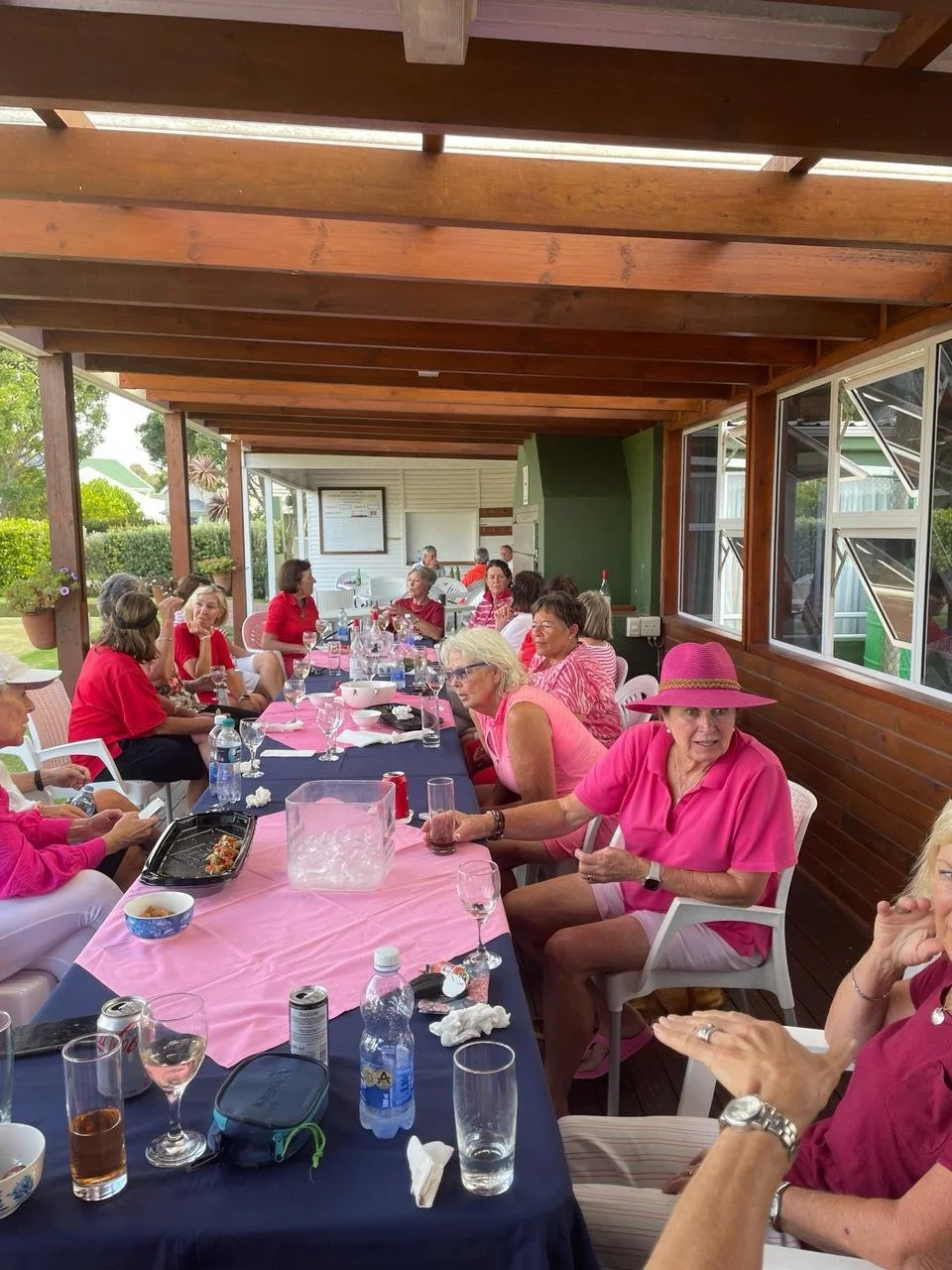 Group of women sitting around a long table on a covered patio, some wearing pink, pink tablecloth, various drinks and dishes on the table, with a windowed wall and greenery outside.