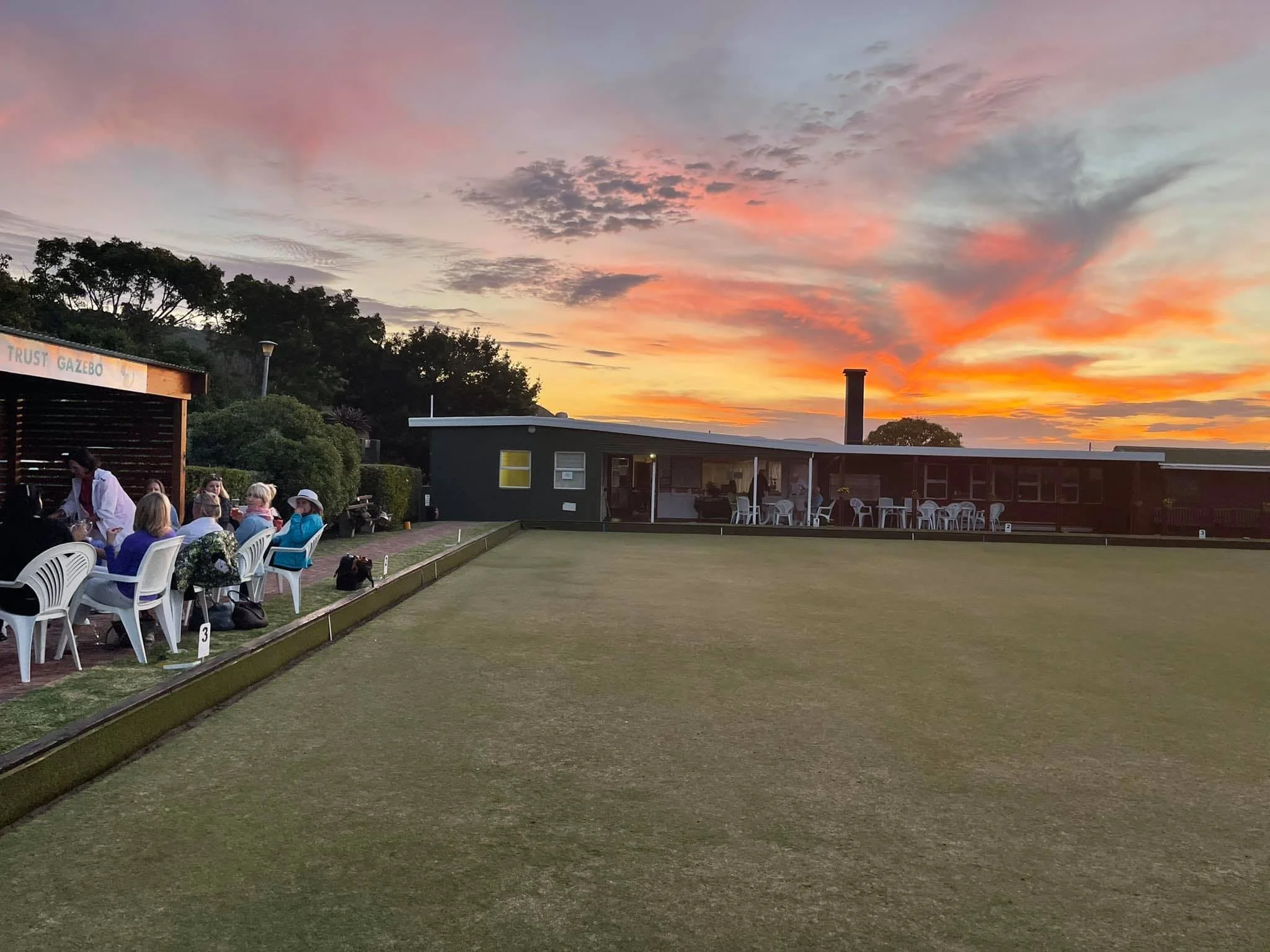 A group of people sitting on white plastic chairs on a patio next to a green lawn at sunset, with a colorful sky and trees in the background.