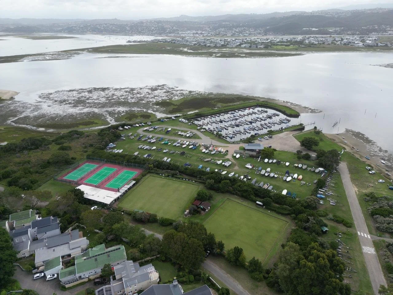 Aerial view of a waterfront park with tennis courts, green fields, and a marina with boats, surrounded by residential houses and water bodies.