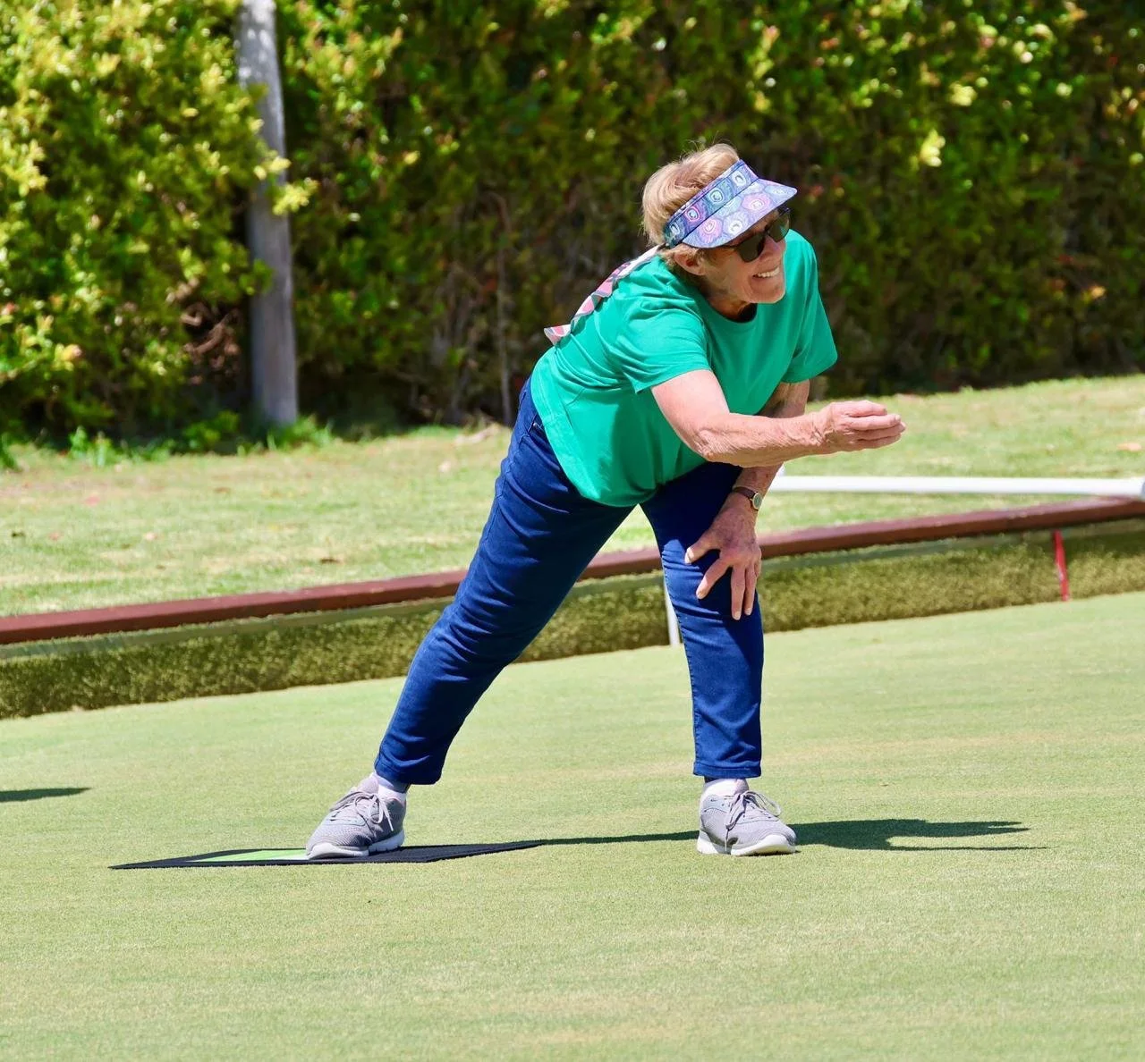 An elderly woman with short gray hair, wearing sunglasses, a colorful visor, a green T-shirt, and blue jeans, is bending forward on a golf course, appearing to line up a putt or to practice putting.