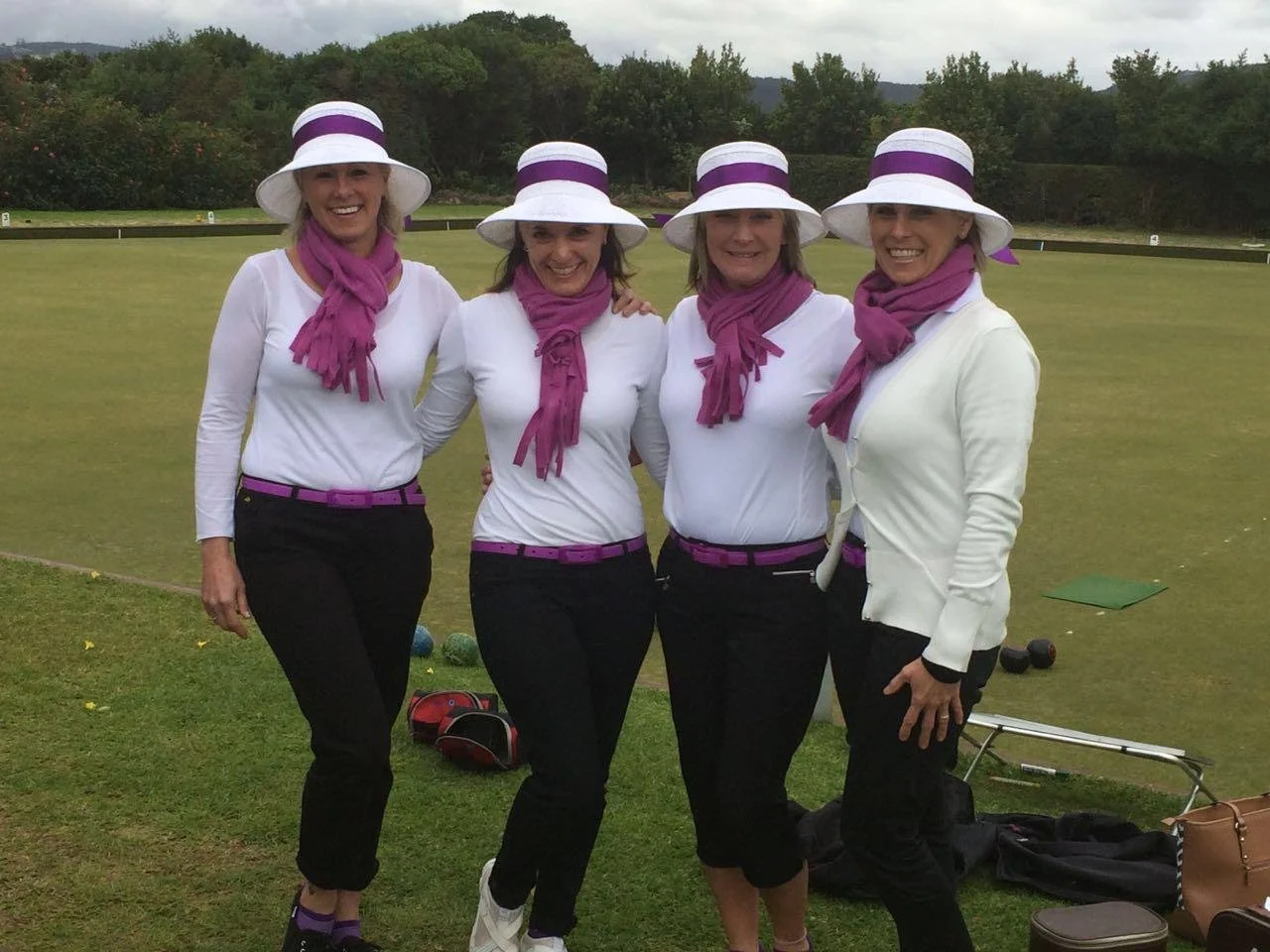 Four women standing together on a golf course, dressed in white shirts, black pants, purple scarves, and white hats with purple bands, smiling at the camera.