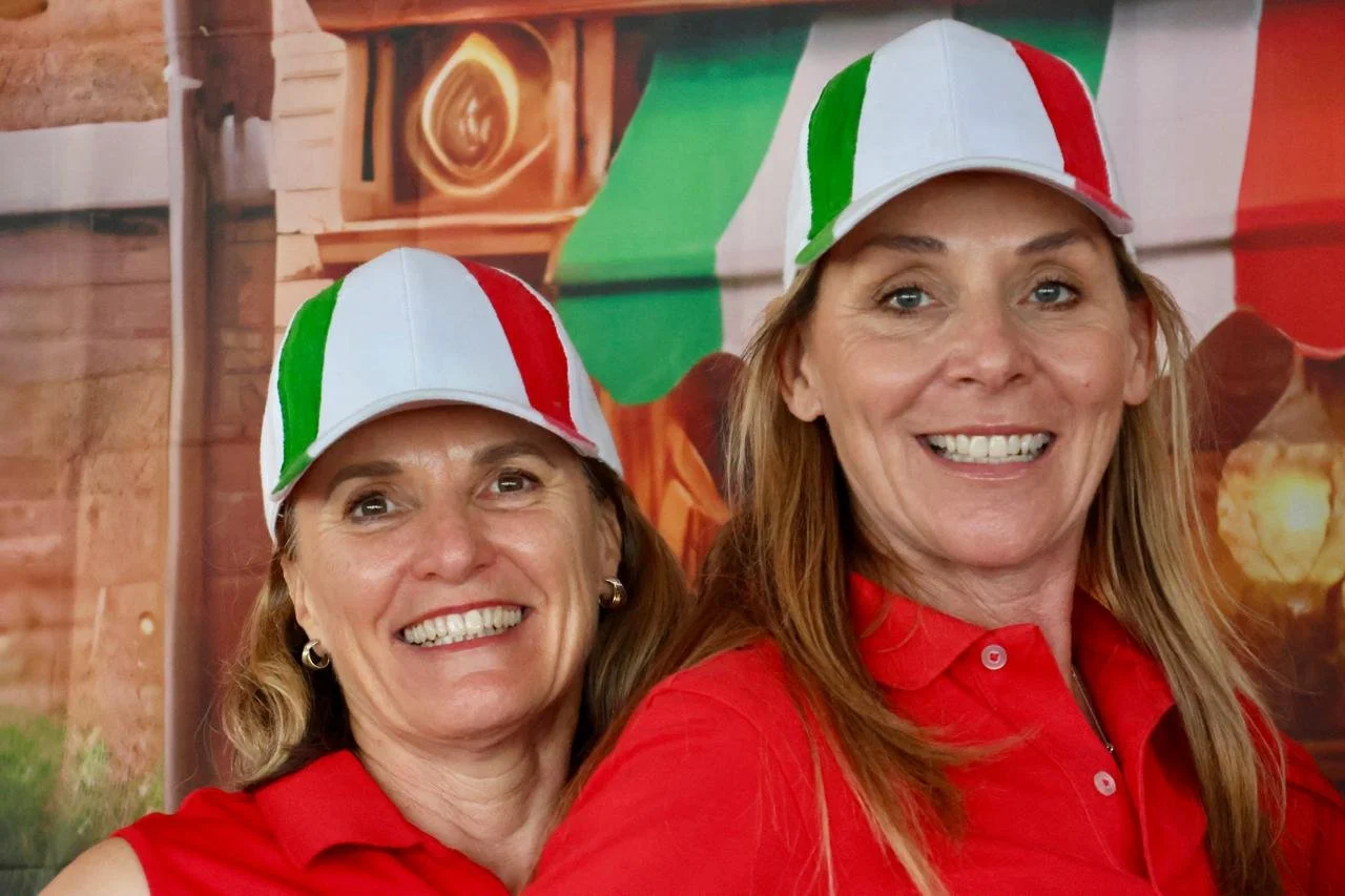 Two women smiling, both wearing white caps with green, white, and red stripes, and red shirts. The background features a banner with Italian flags and food imagery.