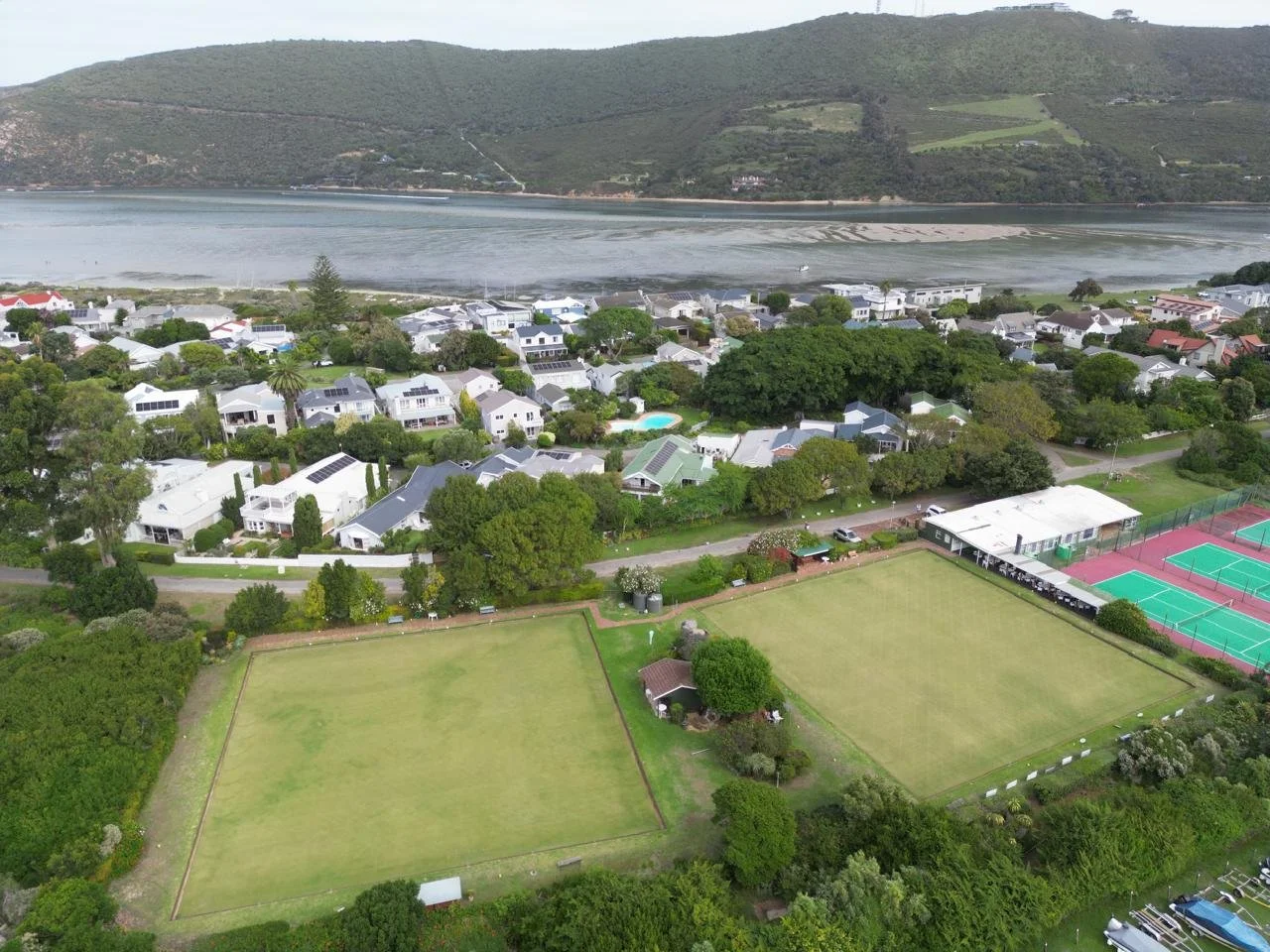 Aerial view of a coastal neighborhood with tennis courts, houses, a small pool, and a river or bay in the background surrounded by hills.