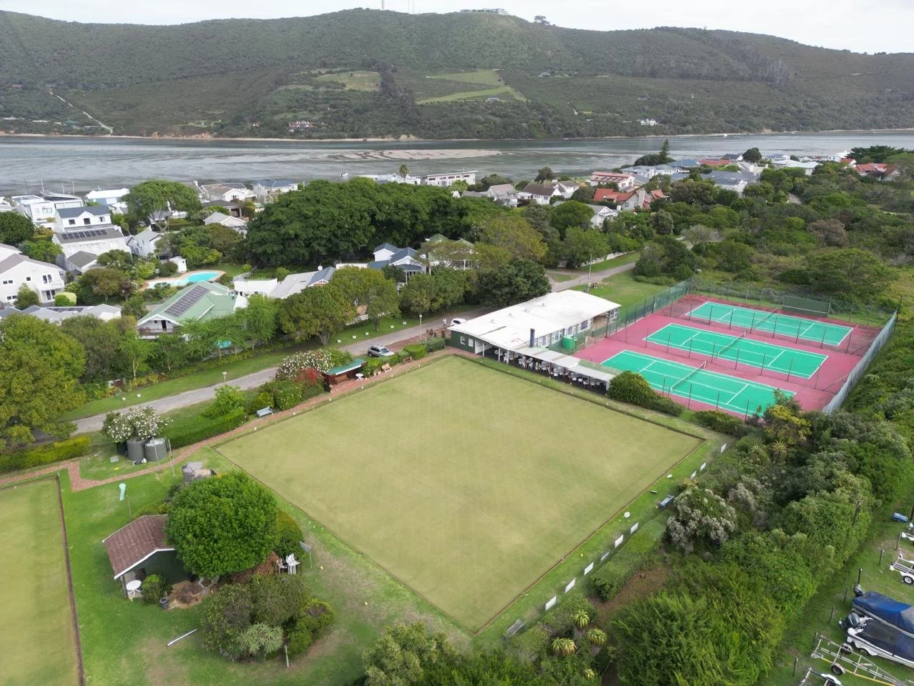 Aerial view of a recreational area with tennis courts, a large grassy field, a clubhouse, and surrounding houses with a river and hillside in the background.
