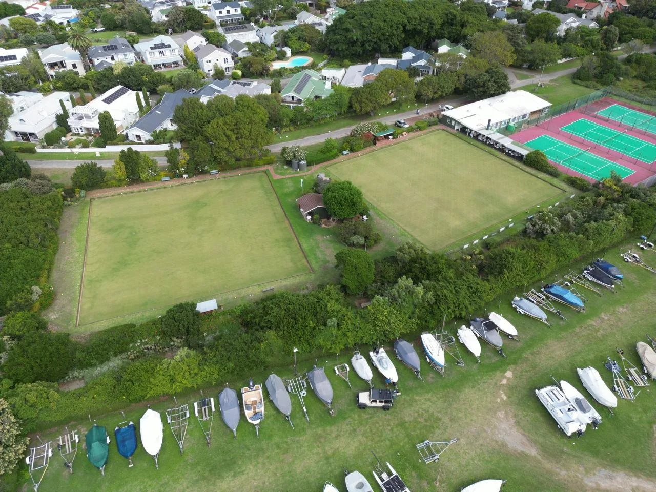 Aerial view of a neighborhood with tennis courts, grassy fields, boats, and residential houses surrounded by trees.