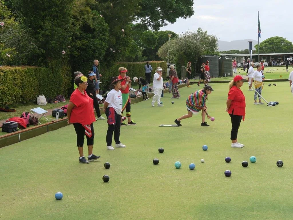 People playing lawn bowls on a green, with some spectators in the background, trees, and a flagpole.