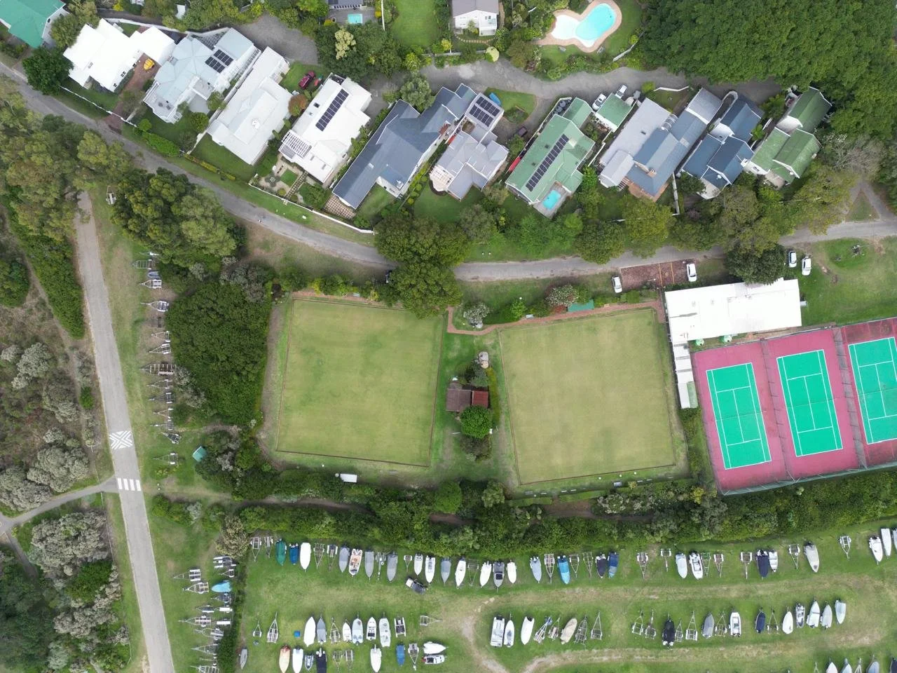 Aerial view of a residential area with houses, tennis courts, grass fields, parking lots, and surrounding trees.