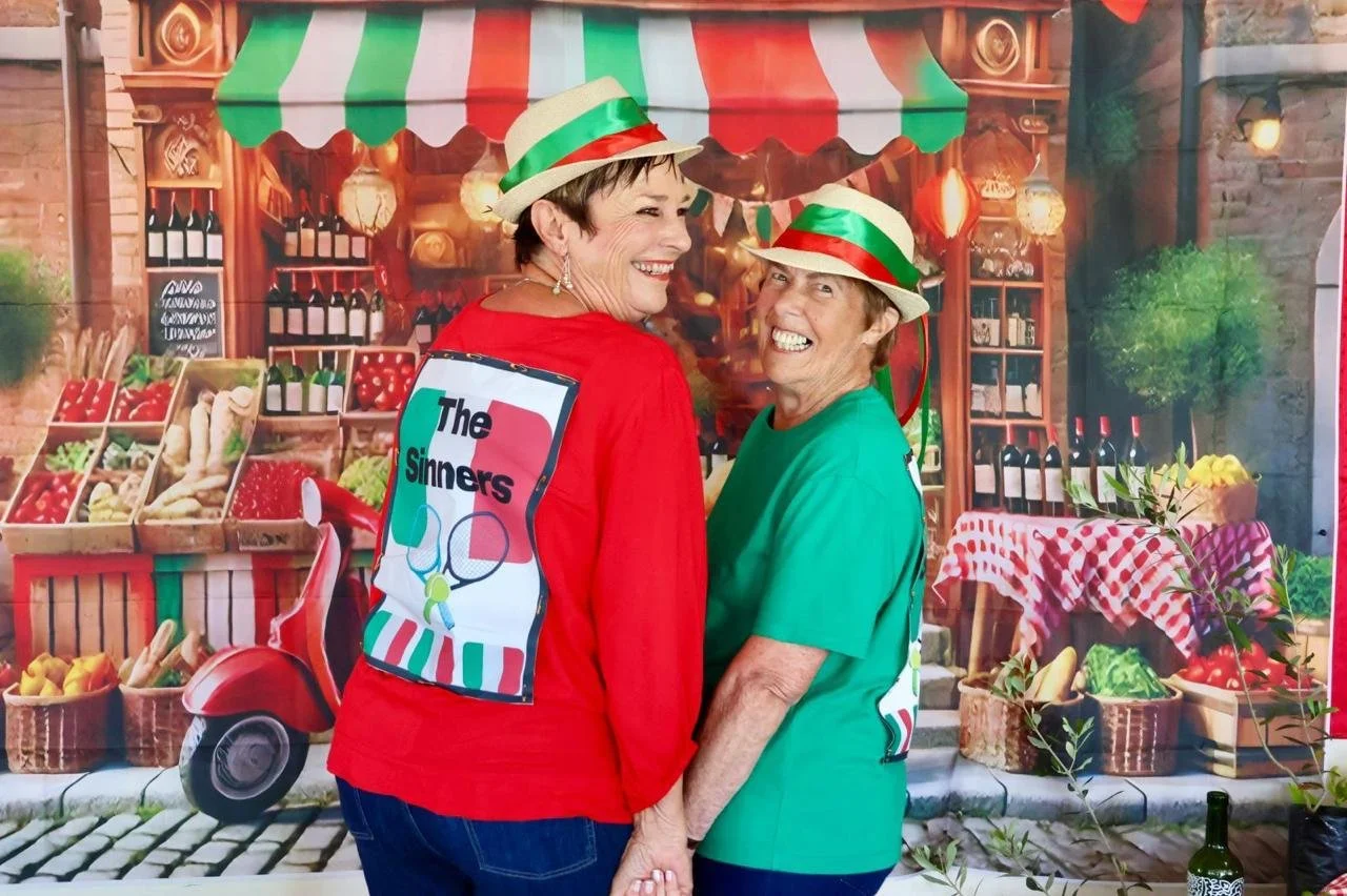 Two women wearing festive hats and colorful shirts, standing in front of a market backdrop, smiling and holding hands.