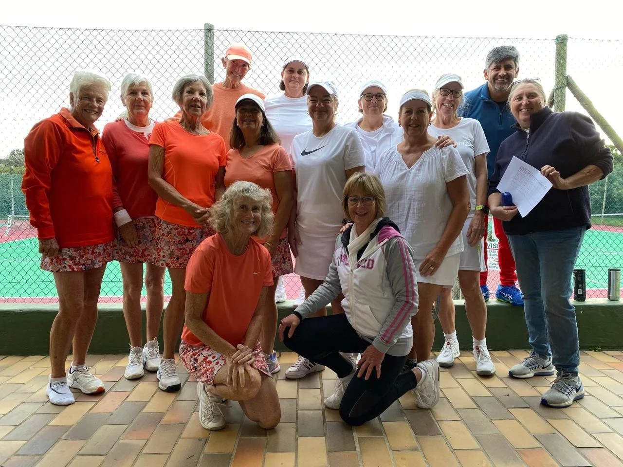 Group of senior women and a man standing and kneeling in front of a tennis court, dressed in sportswear, smiling and posing for a photo.