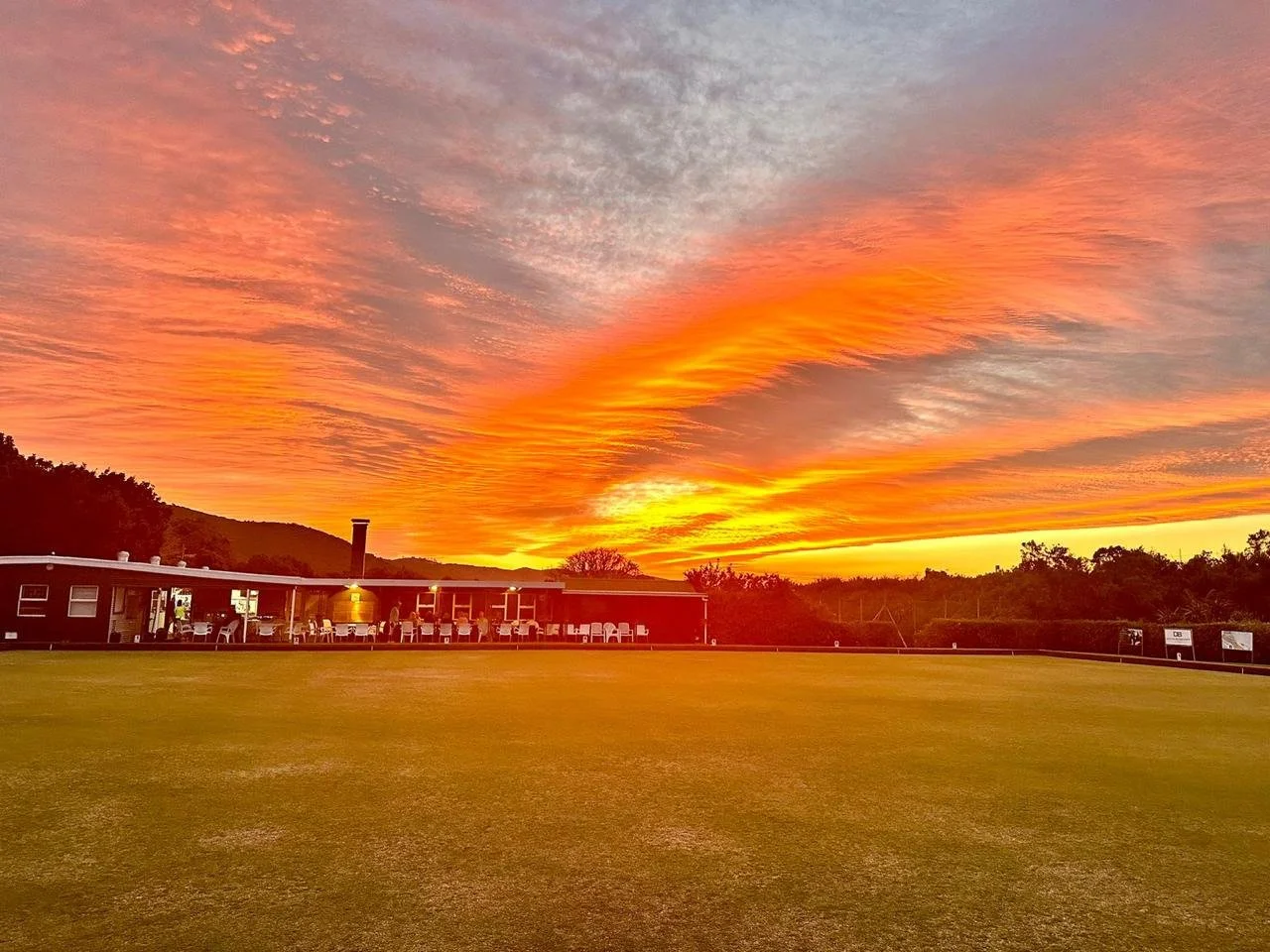 Sunset over a sports field with a clubhouse and distant trees and hills.