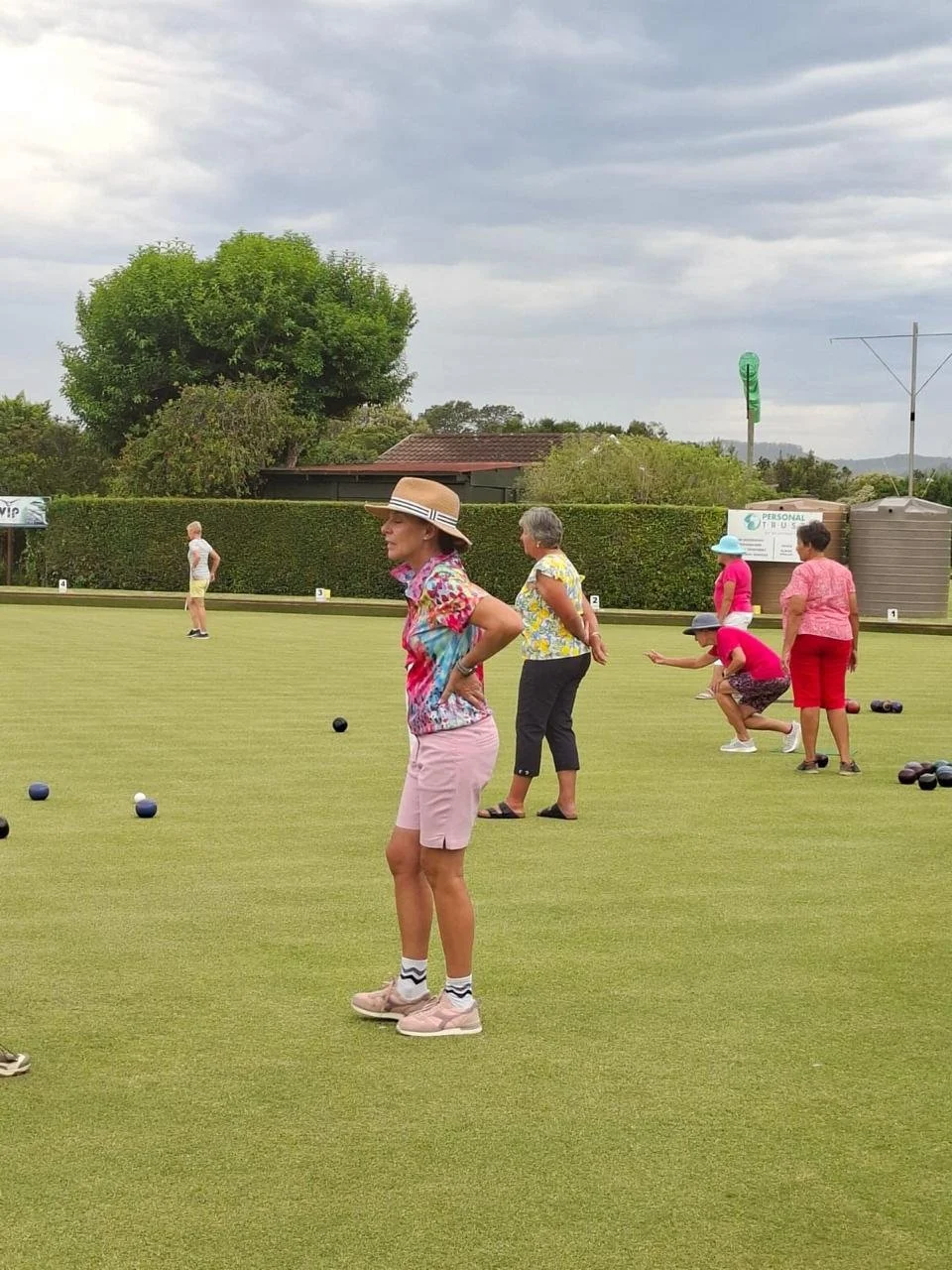 Women playing lawn bowls on a green, dressed in colorful summer clothes and hats, with a cloudy sky overhead.