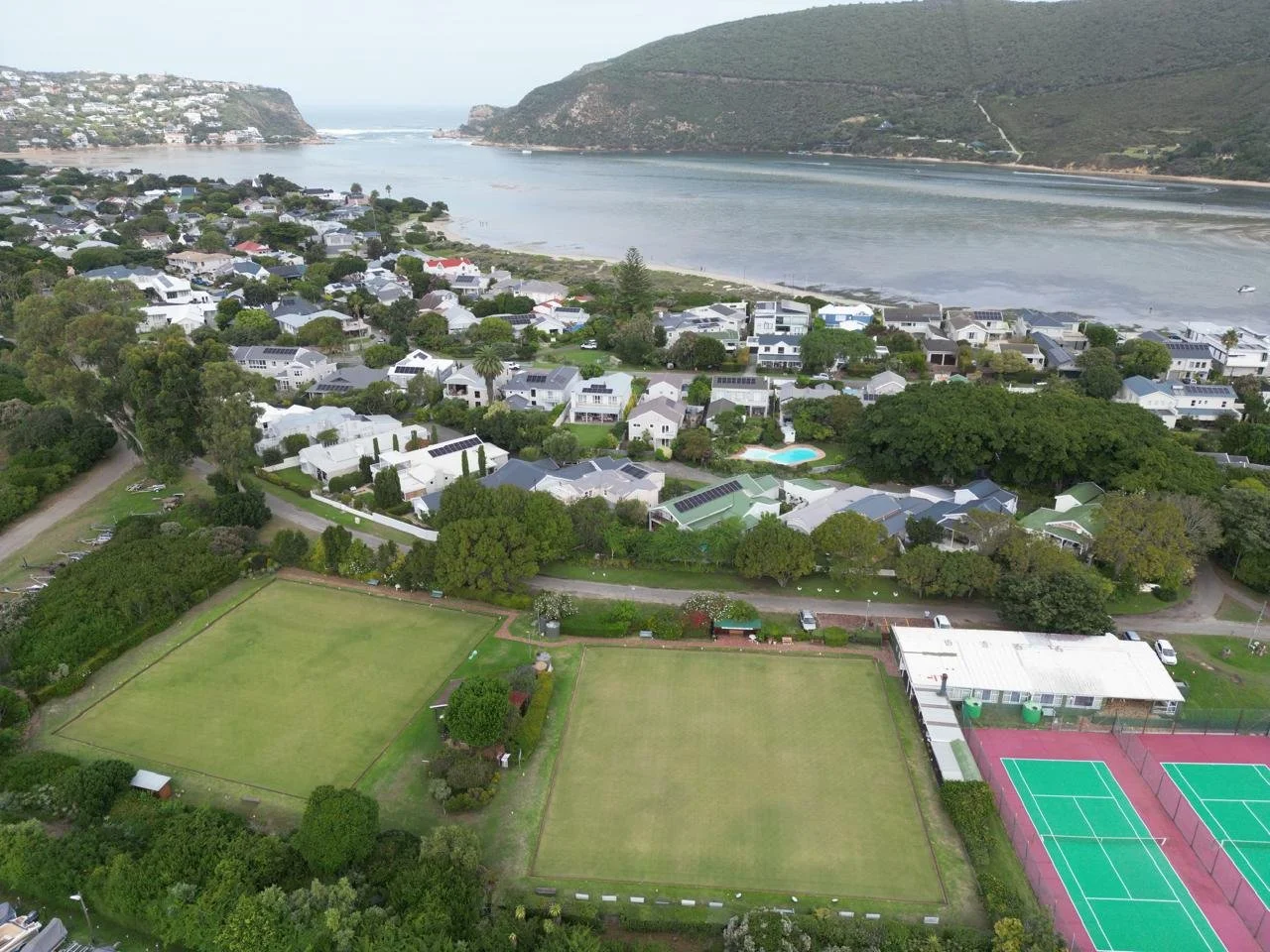 Aerial view of a residential neighborhood near a bay, with houses, tennis courts, a swimming pool, green fields, and surrounding trees and hills in the background.