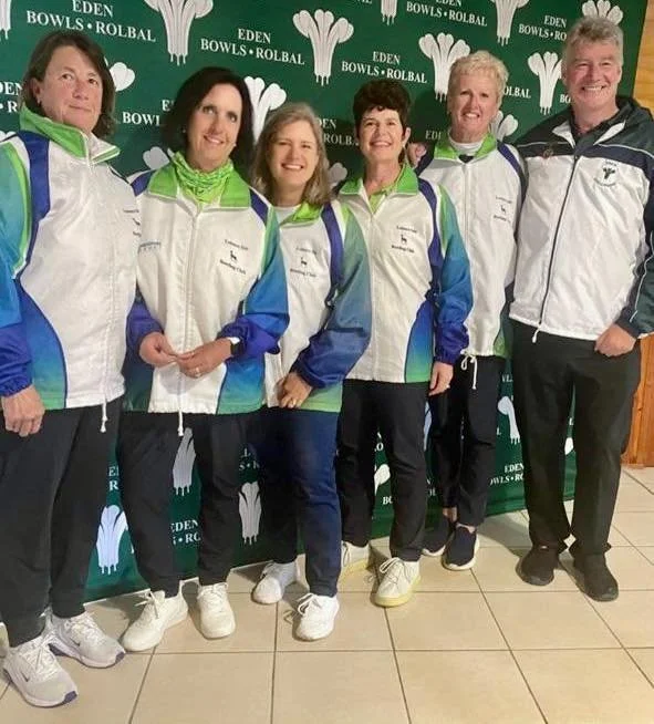 Group of six people wearing matching sports jackets with blue, green, and white colors, standing in front of a green backdrop with the words 'Eden Bowls Roll Ball' and a graphic of a bowling pin.