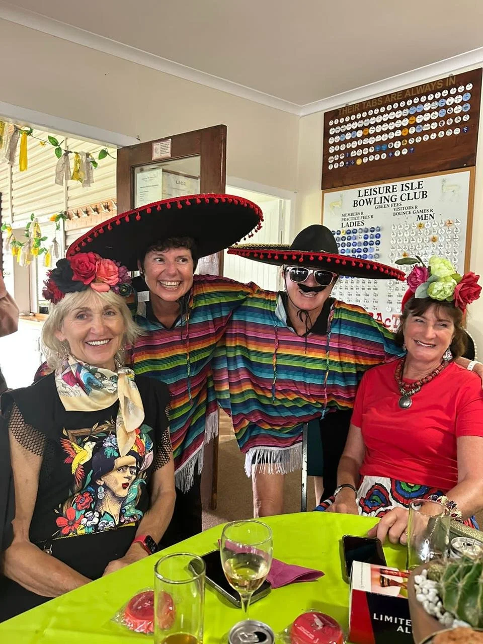 Four women wearing colorful traditional Mexican costumes, including large sombrero hats and floral headpieces, smiling at a celebratory gathering indoors.
