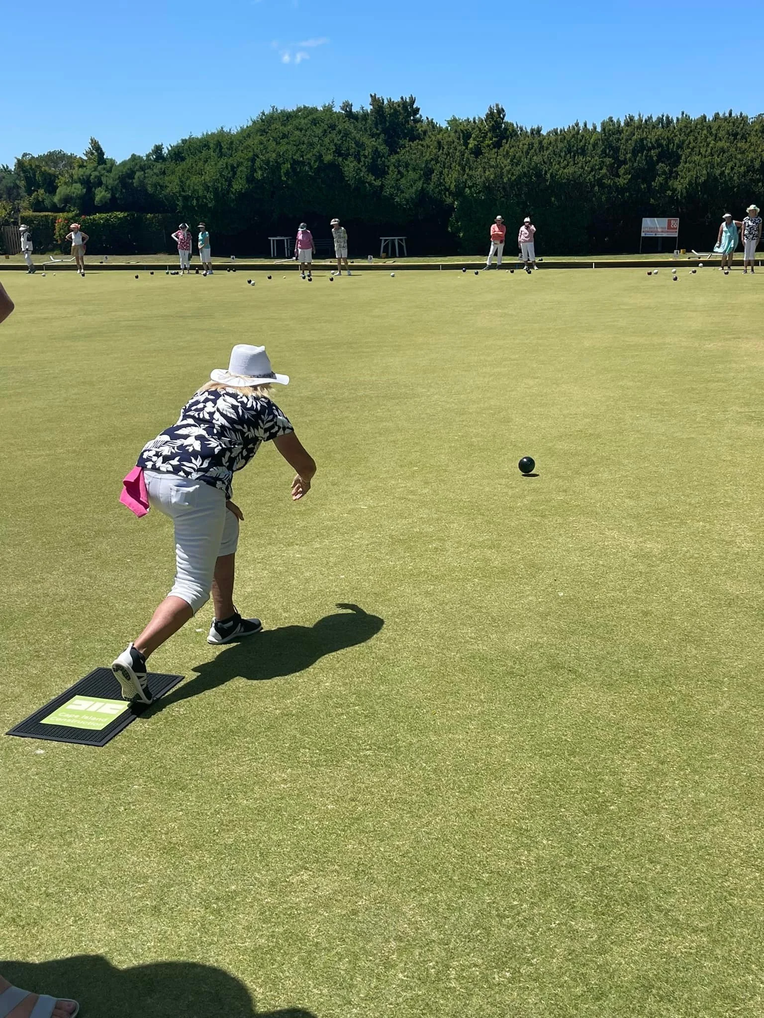 A woman wearing a white hat, black floral shirt, white shorts, and sneakers is playing lawn bowls on a green field, with several other people in the background.