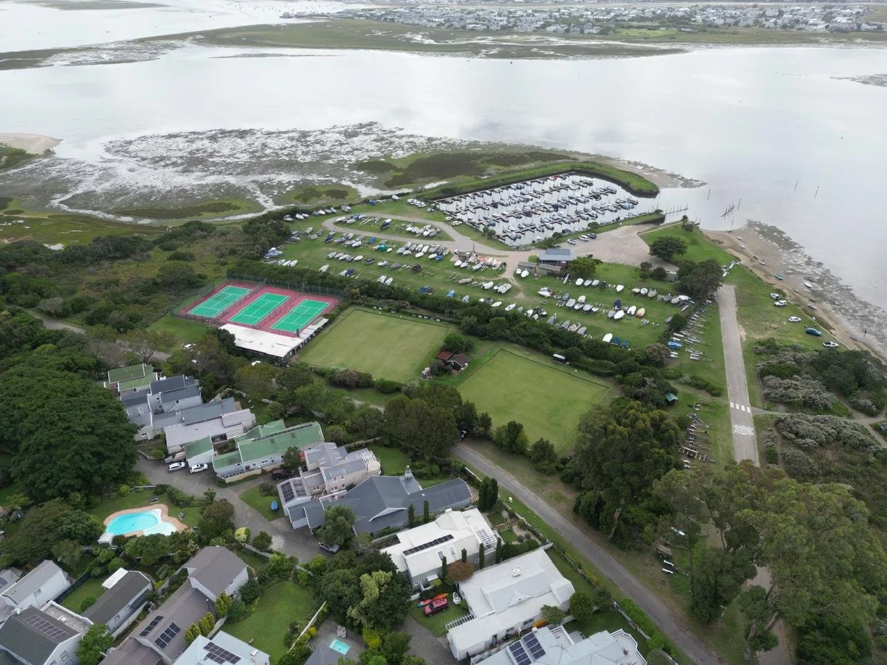 Aerial view of a waterfront community with houses, tennis courts, a swimming pool, green fields, a parking lot filled with boats, and a large body of water in the background.