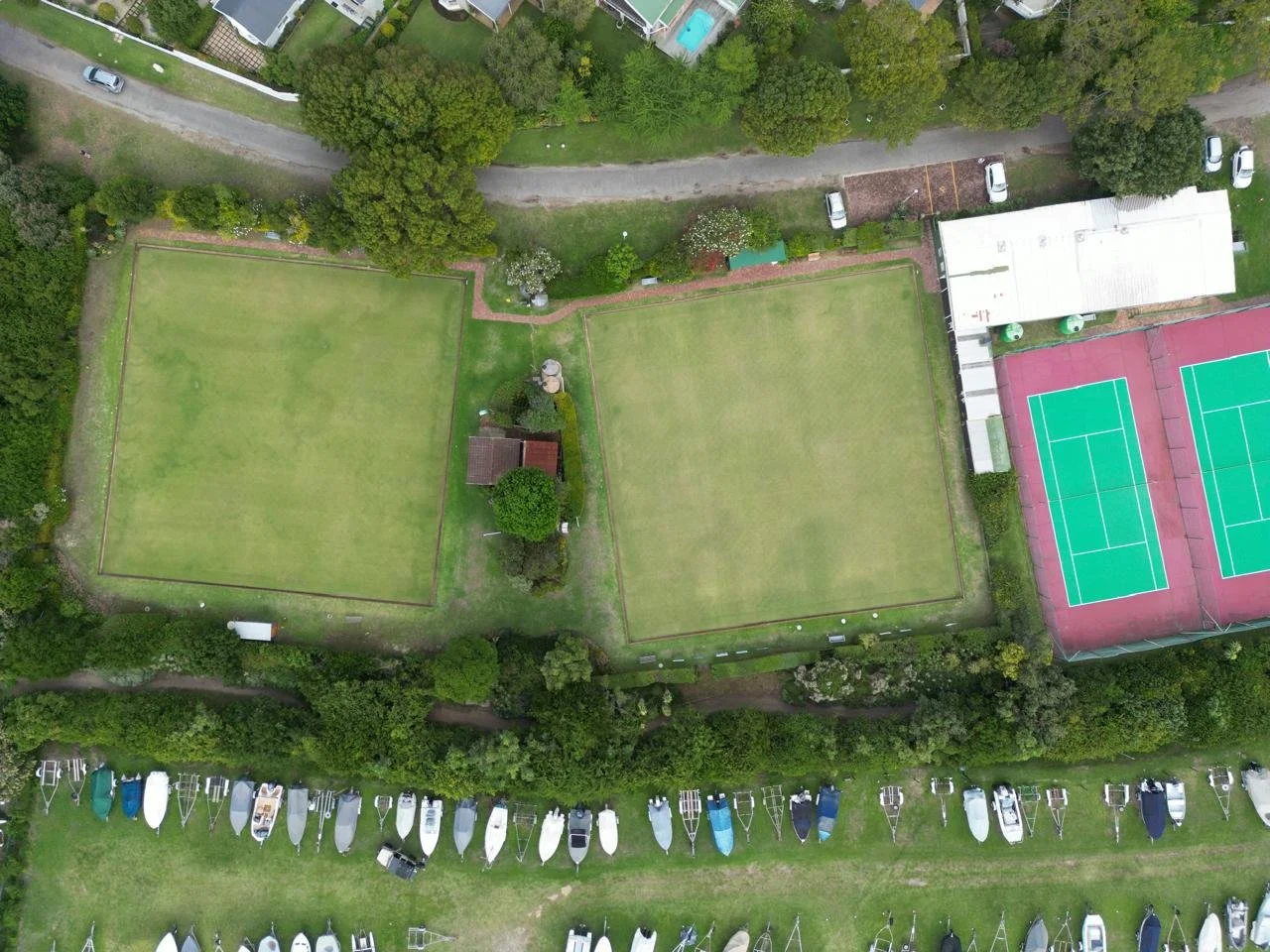 An aerial view of two green tennis courts, a building, parked boats, and surrounding trees and pathways.