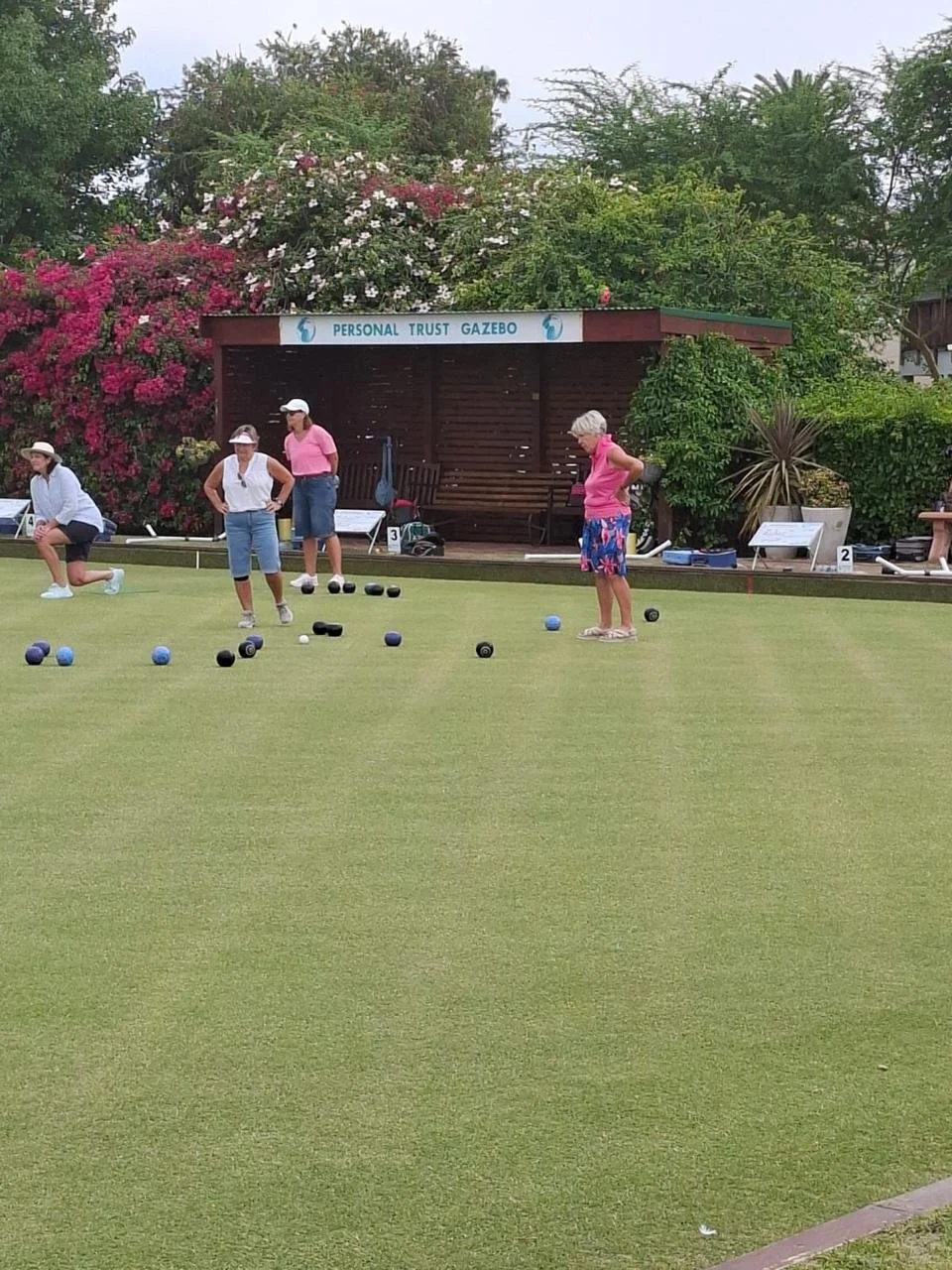 Women playing lawn bowls on a green during daytime, with a wooden gazebo labeled 'Personal Trust Gazebo' and lush flowered bushes in the background.