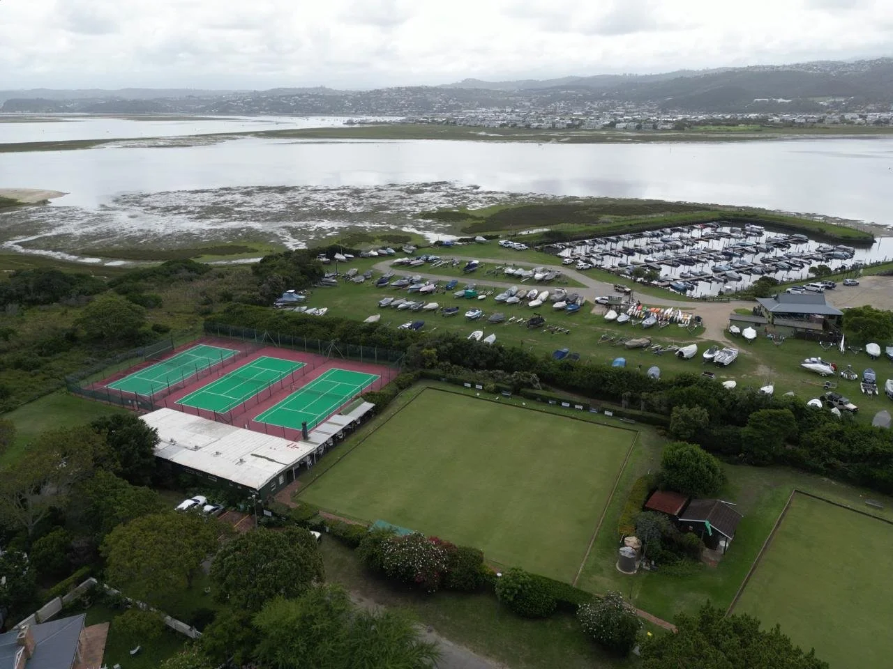 Aerial view of sports complex with tennis courts, grass fields, parking lot, boats, marina, and water body in the background.