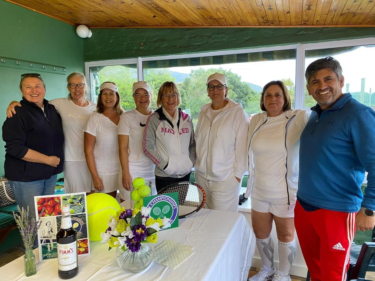 Group of eight people smiling, standing indoors with tennis equipment and decorative items on a table in front of them, windows showing trees and mountains outside.