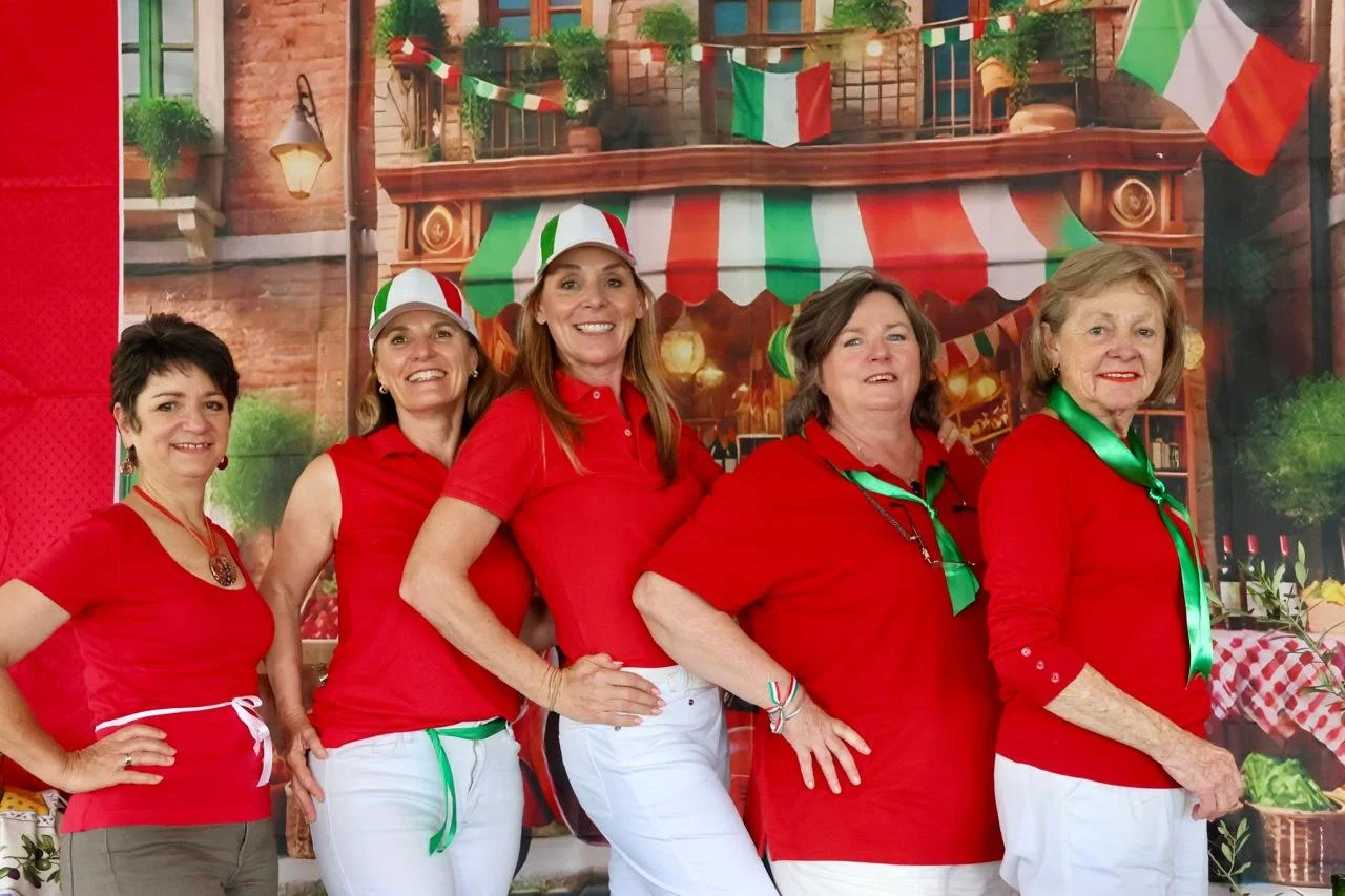 Group of five women dressed in red tops with green, white, and red accessories, standing in front of Italian-themed backdrop.
