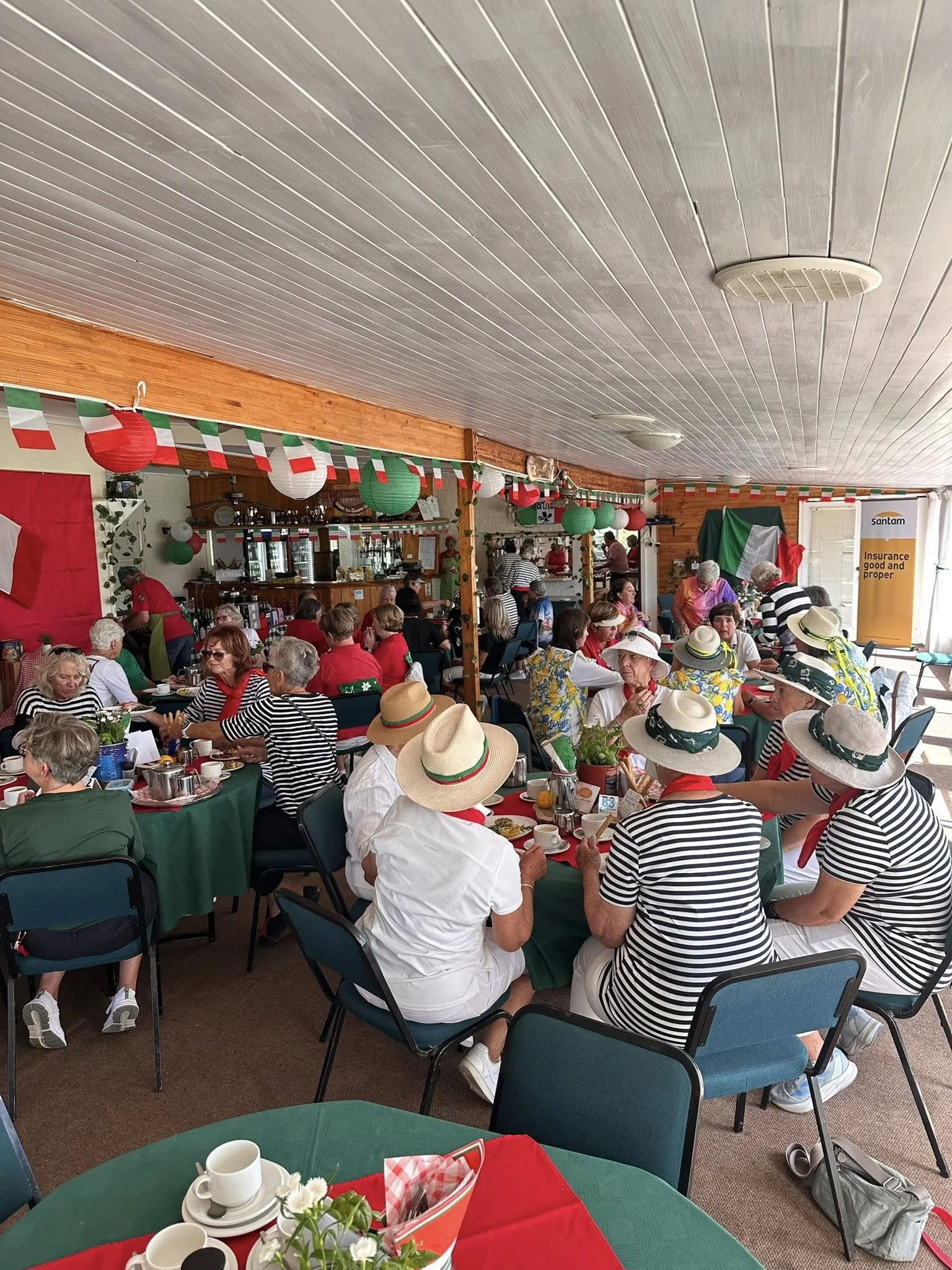 A large group of elderly women gathered indoors for a celebration, many wearing striped shirts, hats, and red scarves. The room is decorated with green, red, and white paper lanterns and banners, with an Italian flag and a banner with 