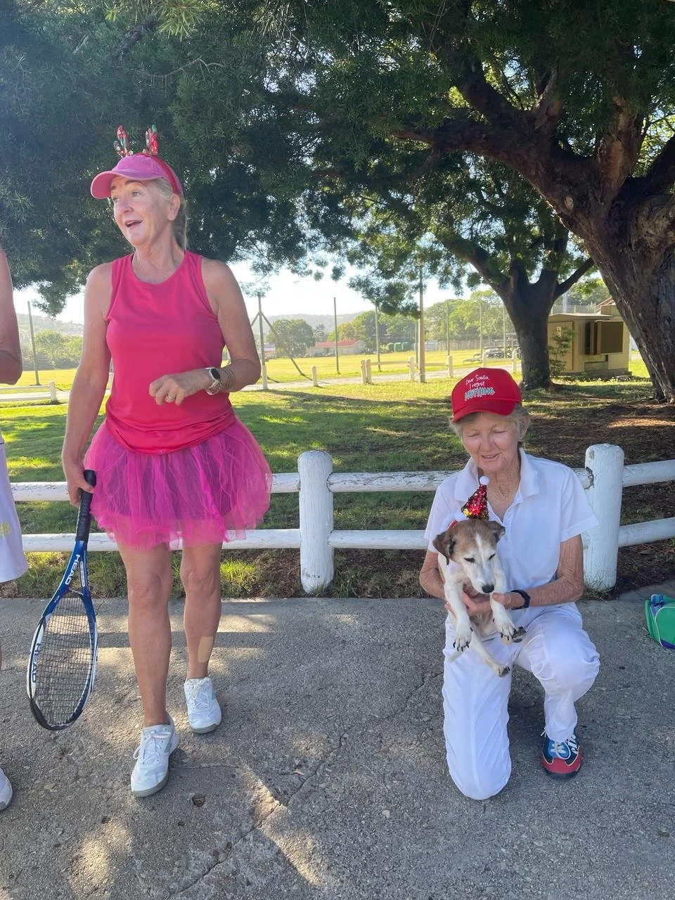 Two elderly women outdoors under a large tree. One woman is standing and wearing pink athletic clothing and a pink cap with antler decorations, holding a tennis racket. The other woman is sitting and dressed in white with a red cap, holding a small d