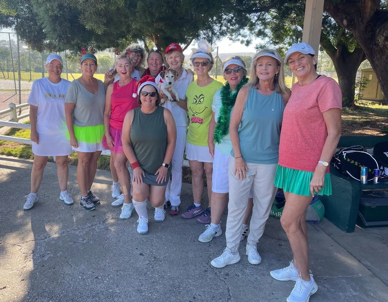 A group of women and a dog gathered outdoors, some women wearing tennis outfits and hats, with festive accessories like tinsel and a Santa hat, under a large tree on a sunny day.