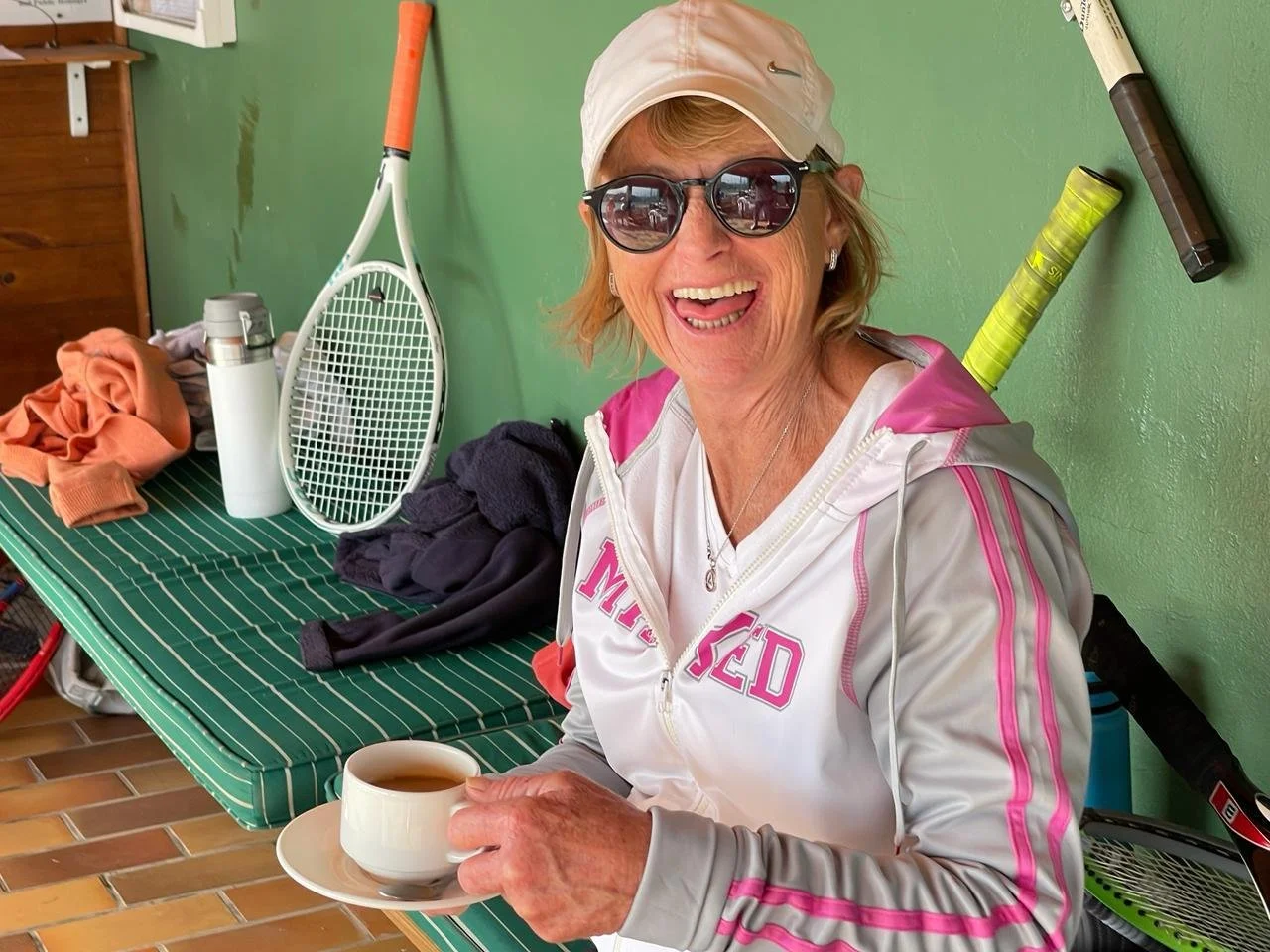 Smiling woman wearing sunglasses, a baseball cap, and a pink and white zip-up jacket, holding a cup of tea or coffee, sitting at a green-striped table with tennis rackets and clothes in the background.