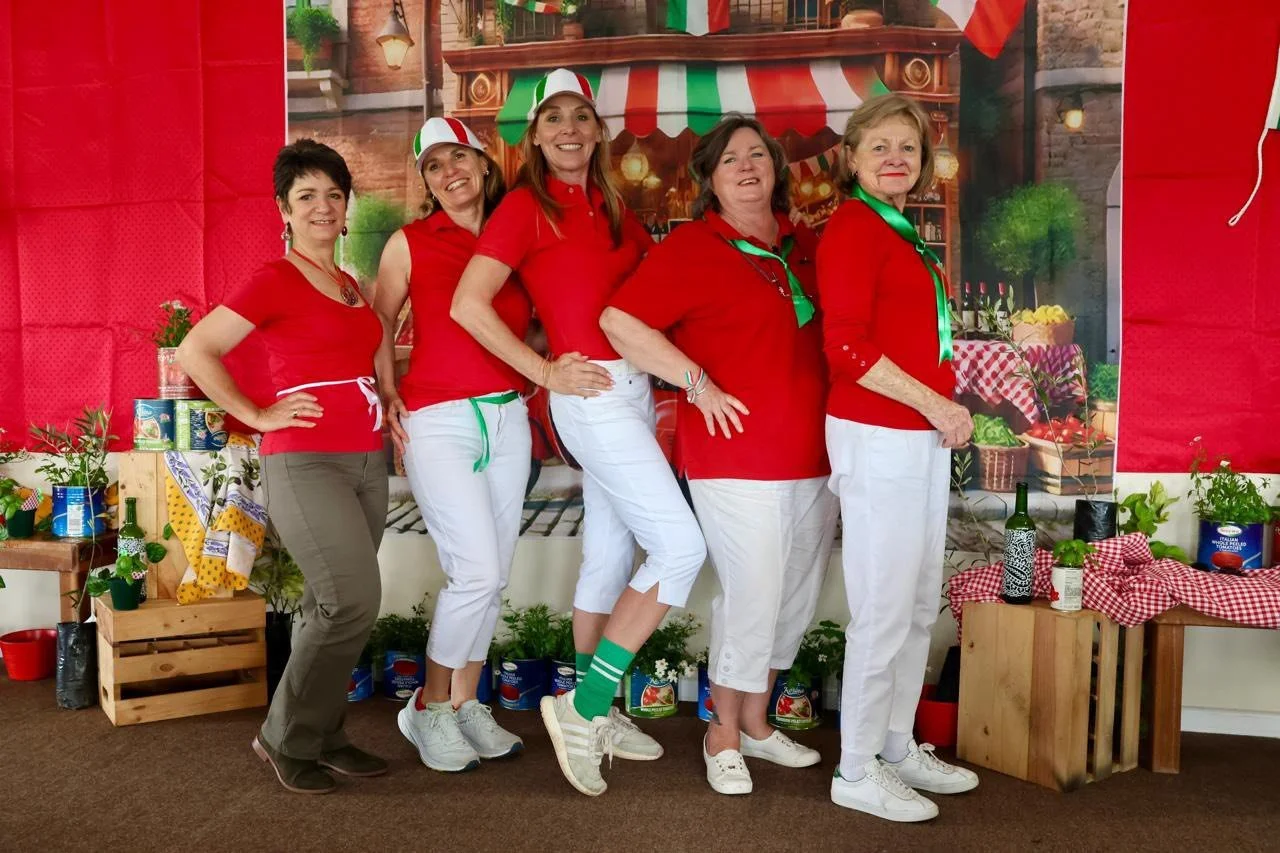 Five women dressed in red and white, standing together indoors in front of an Italian-themed backdrop, smiling at the camera. They are wearing casual clothing with some hats and scarves, with some having green accents. The background features a pictu