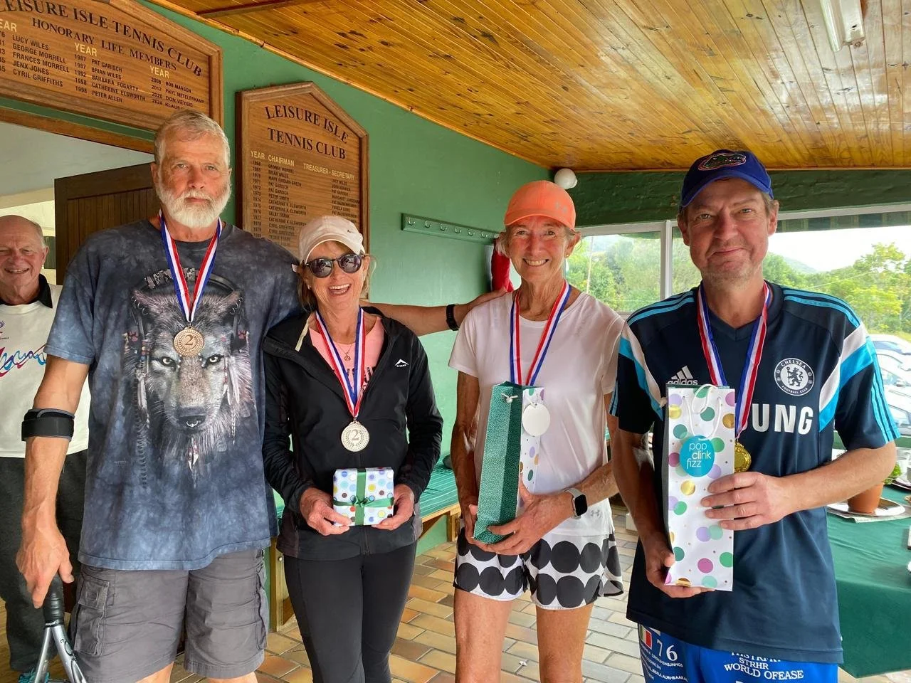Four older adults wearing medals around their necks, standing together in a room with green and wooden walls, celebrating their achievement. They are holding wrapped gifts and smiling. A man in a lion graphic T-shirt is on the left, a woman in sungla