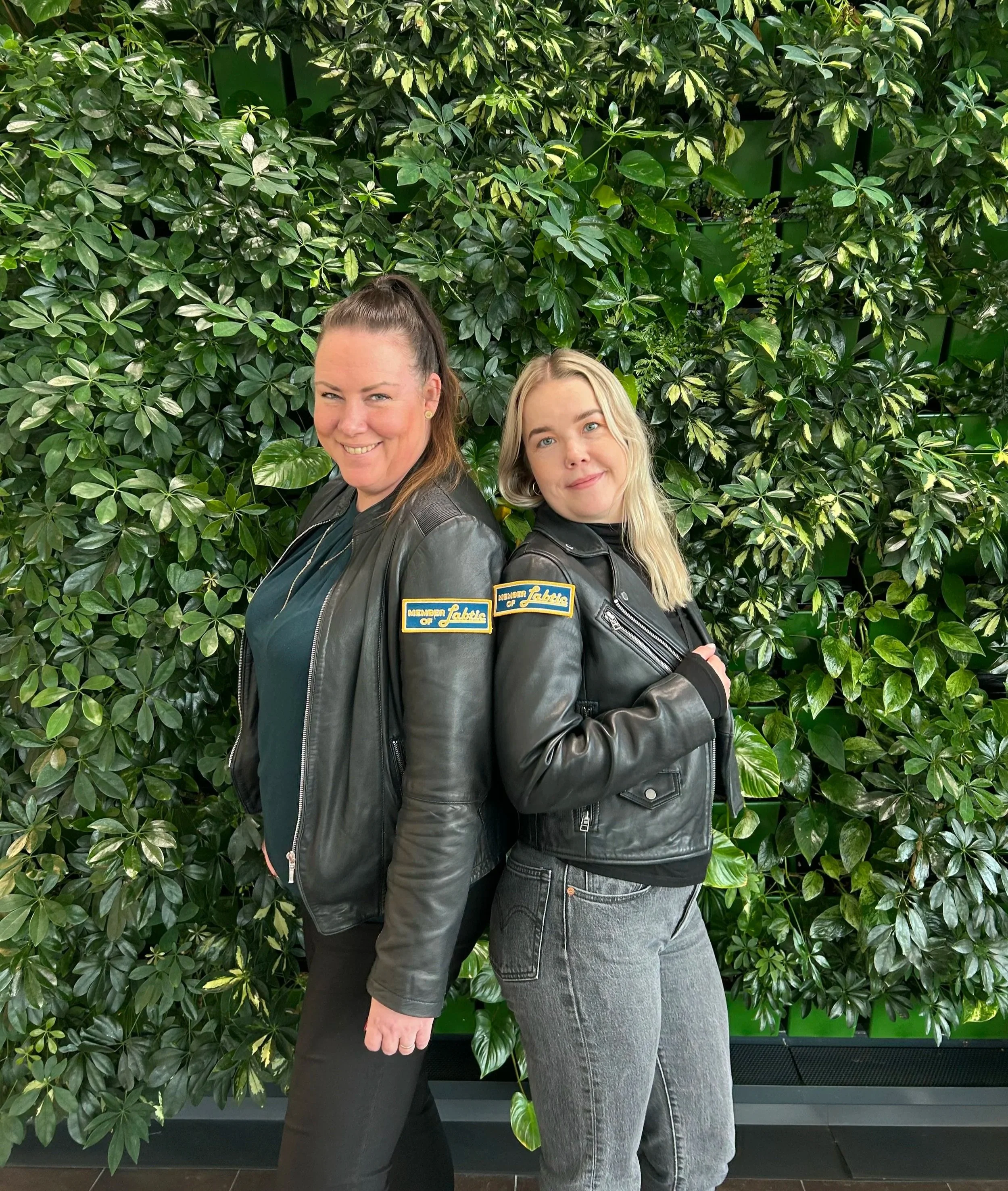 Kaksi naista seisoo selin toisiinsa ja hymyilevät, taustalla vihreitä lehtiä.