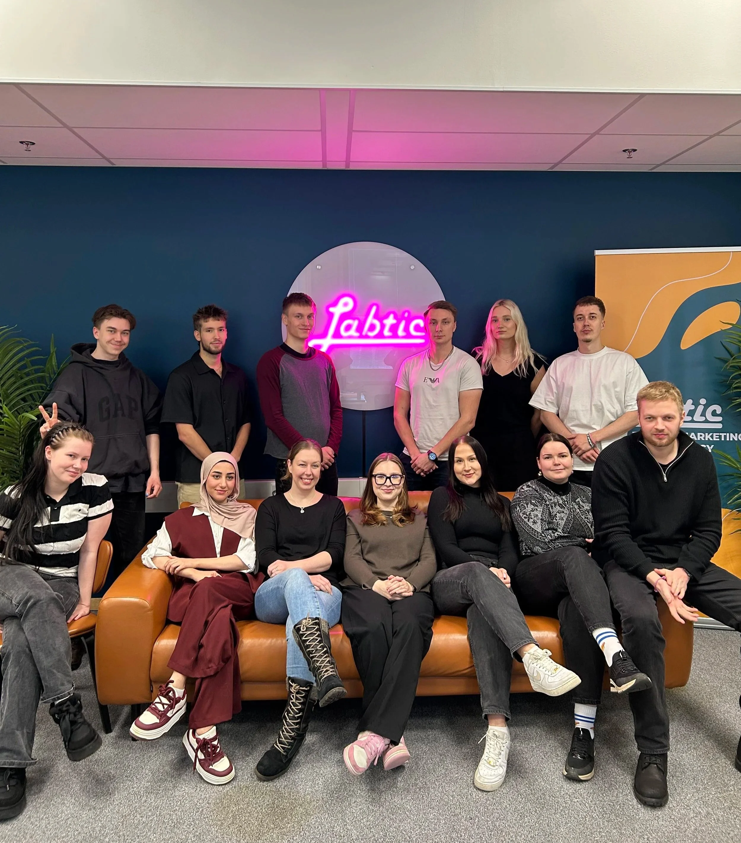 Kuva ryhmästä nuoria ihmisiä istumassa ja seisomassa sisätiloissa, taustalla neonvalomainos lukee 'Labtic'.