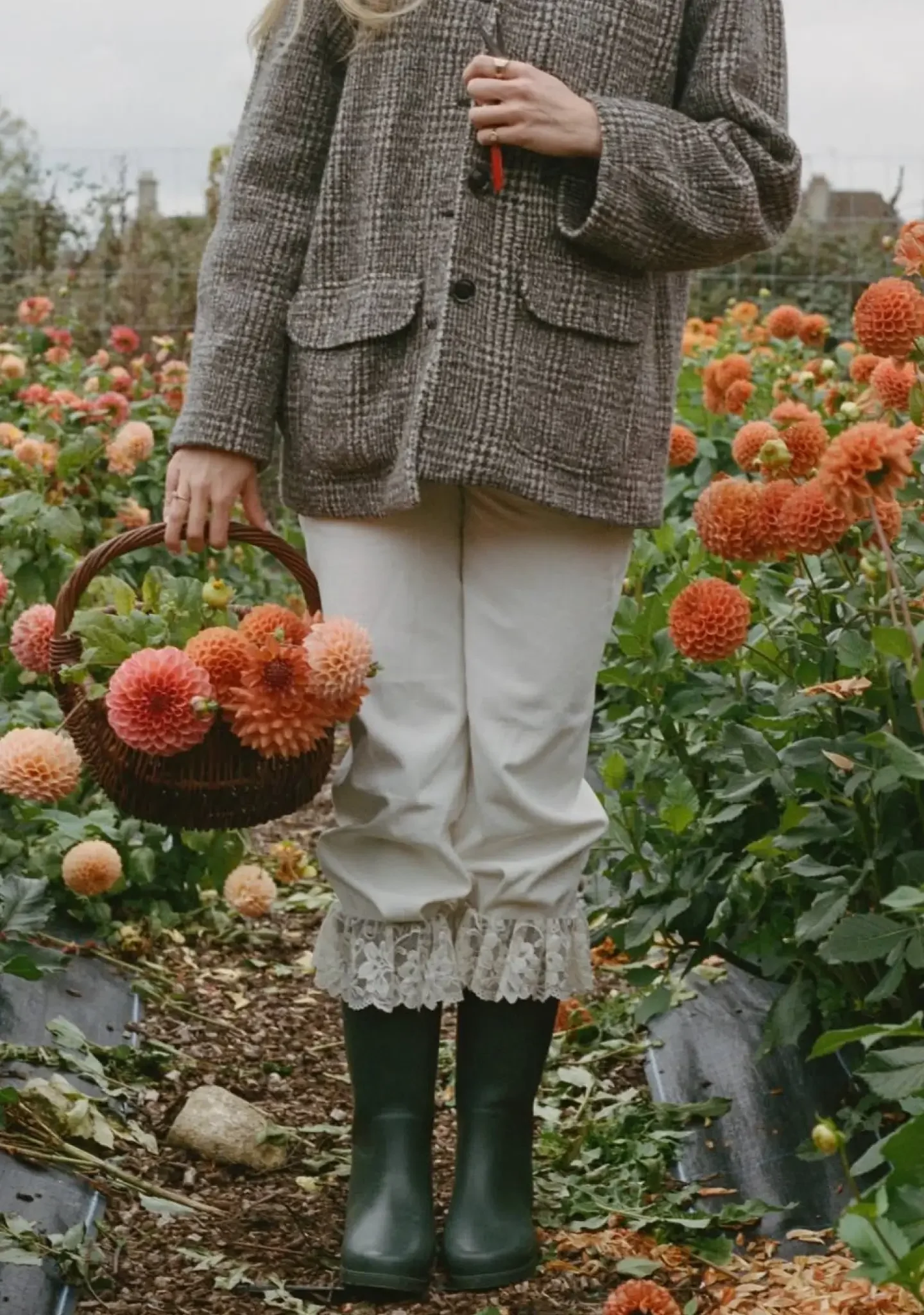 Person standing in a flower field holding a basket of orange dahlias, wearing a brown checked blazer, cream-colored pants with lace trim at the hem, and green rubber boots.