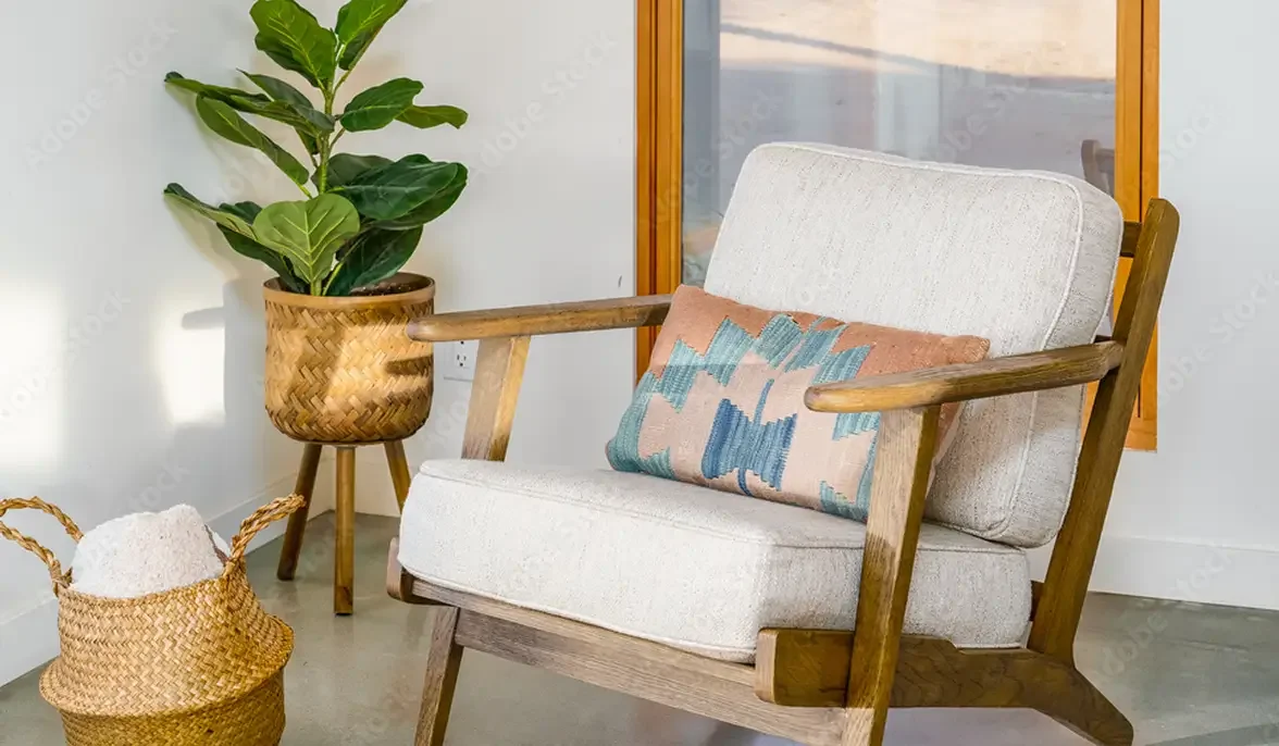 Living room corner with a white upholstered armchair, colorful throw pillow, potted fiddle leaf fig plant in a woven basket, and basket with blanket next to a window with wooden frame