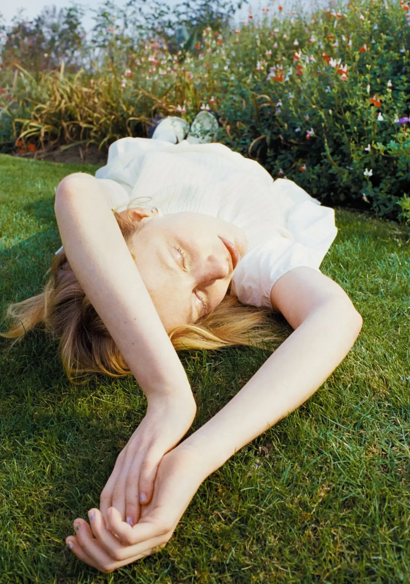 A woman with long red hair lying on green grass with her eyes closed, wearing a white dress, surrounded by blooming flowers.