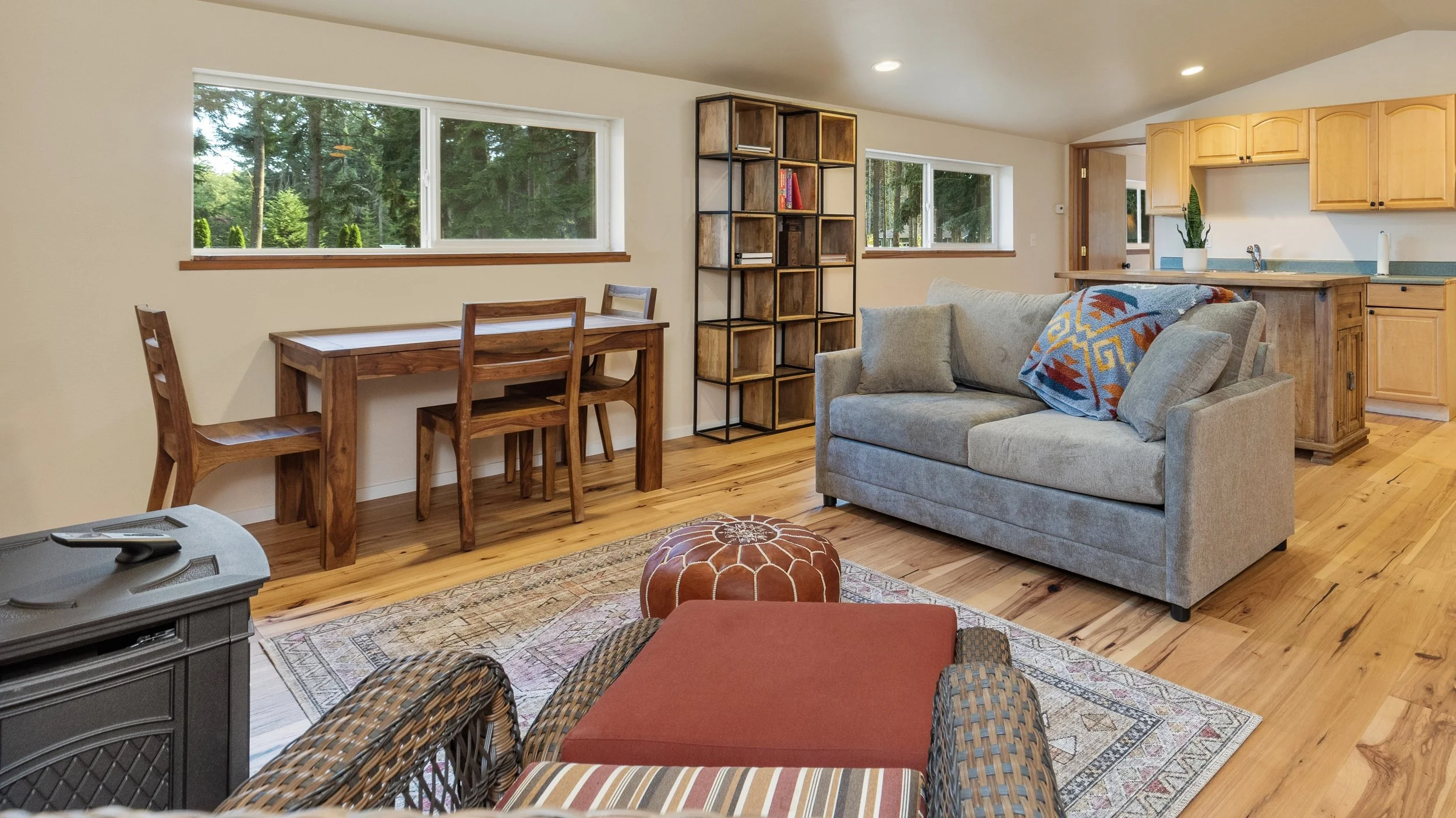 Living room with wooden floor, gray sofa with patterned blanket, wicker chair, wooden dining table with two chairs, bookshelf, window view of trees, kitchen area with wooden cabinets and green countertop, and rug.