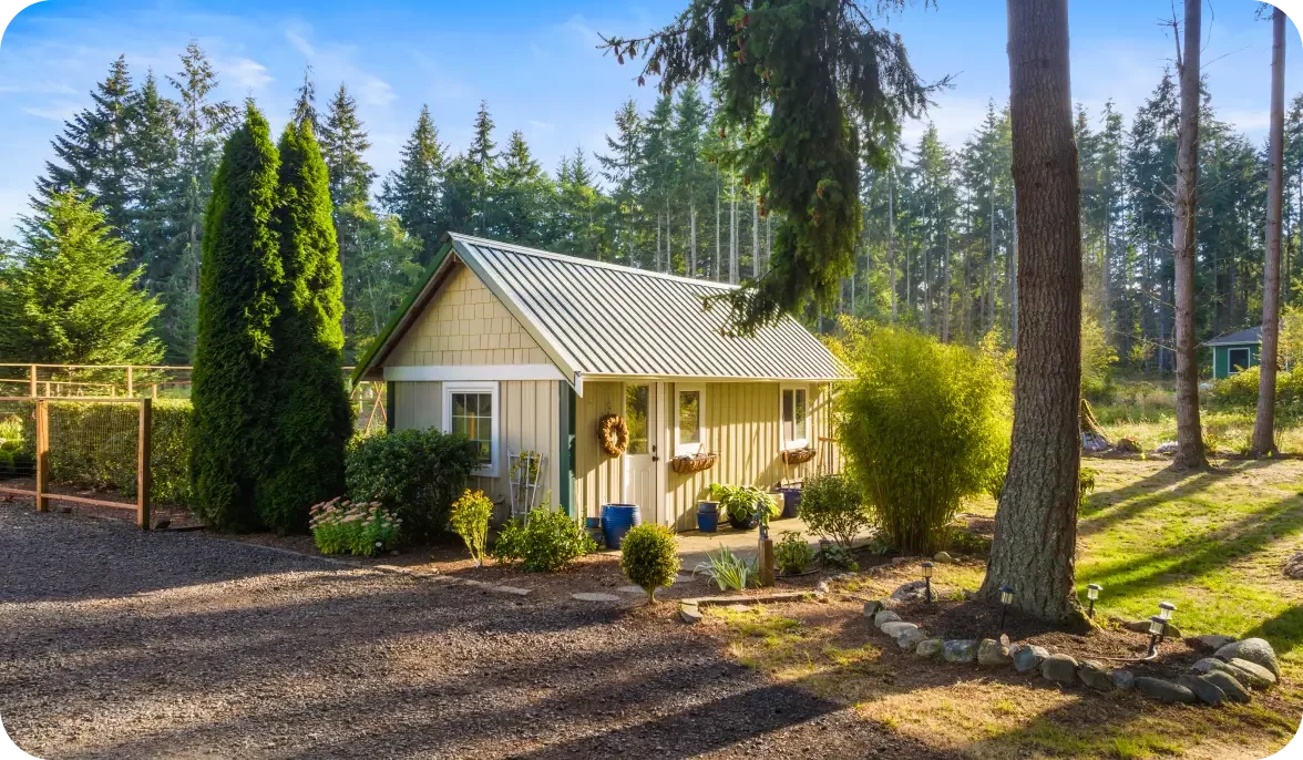 A small house with beige siding and a metal roof, surrounded by trees and shrubs, with a gravel driveway and garden decorations.