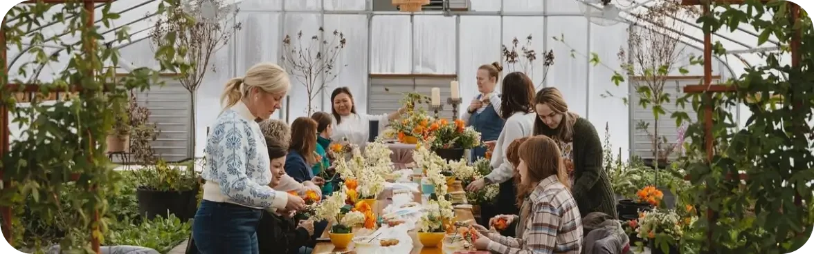 People gathered around a long table in a greenhouse, decorating floral arrangements.