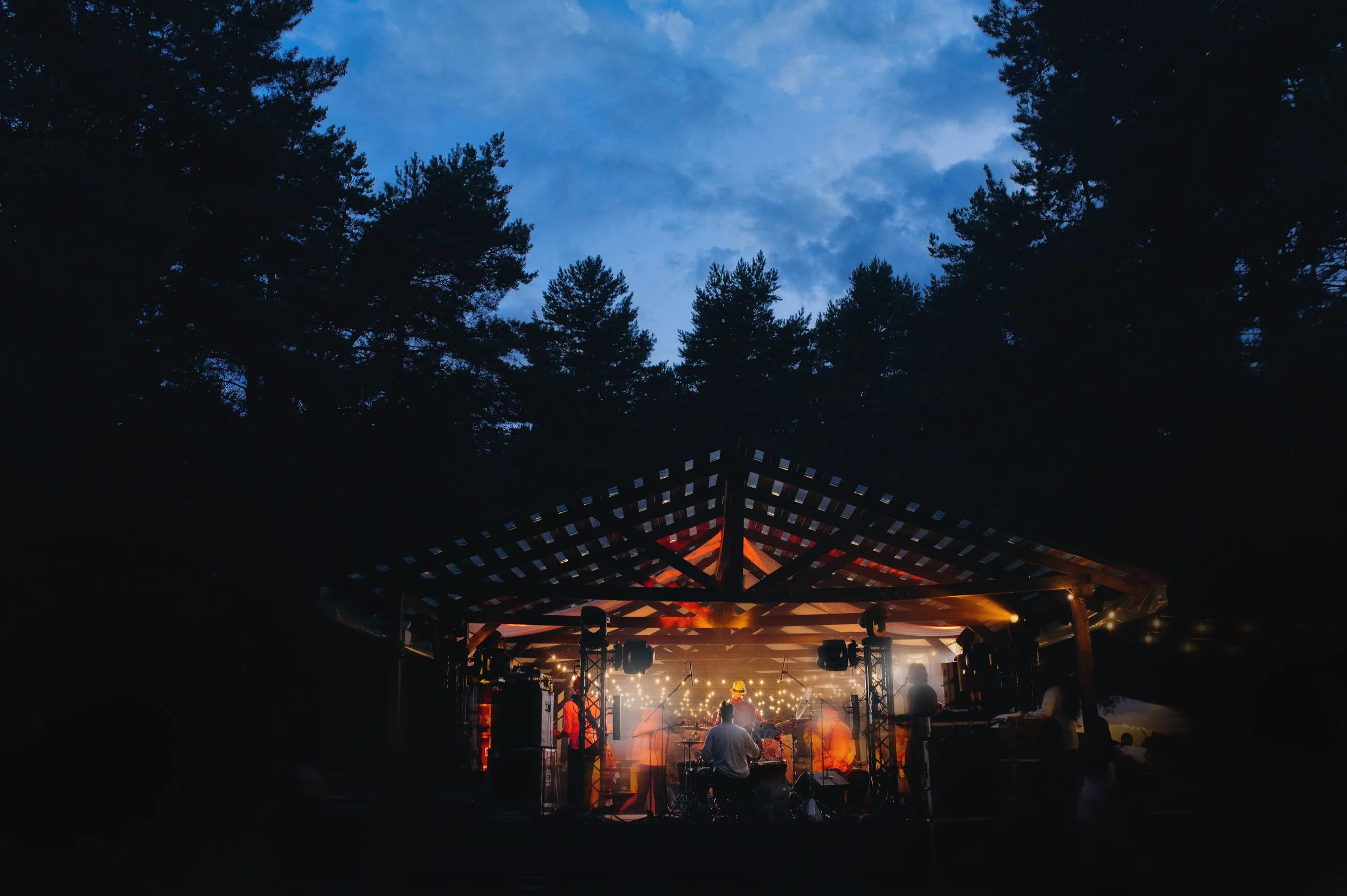 A live outdoor music performance on a stage at dusk, surrounded by trees with a dark blue sky overhead. The stage is lit with warm lights and multiple musicians are playing instruments.