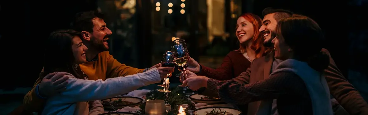 Group of six friends enjoying a toast at a dinner party in a warmly lit restaurant or outdoor setting.