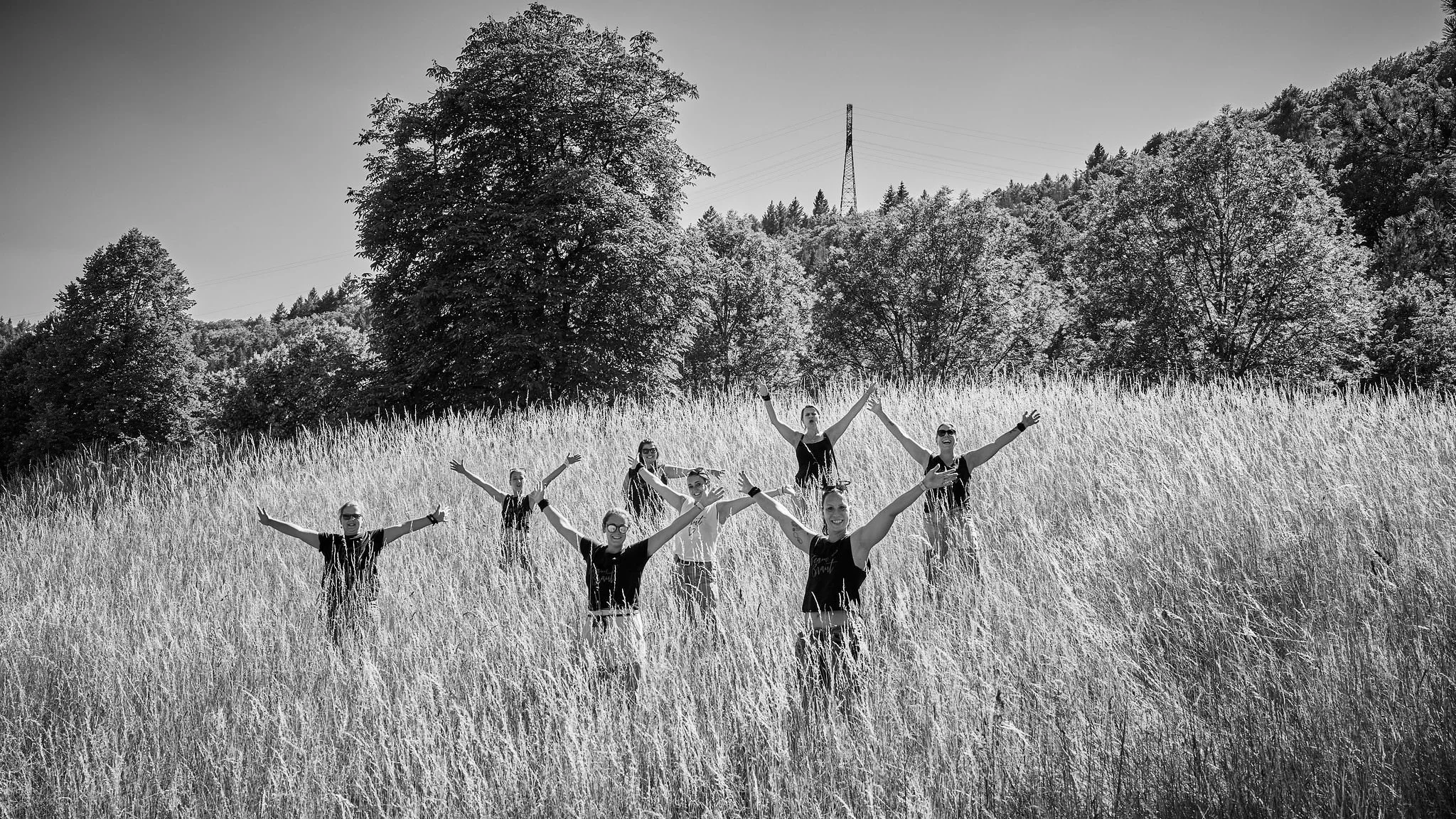 A group of people standing in tall grass, smiling and celebrating outdoors in a field with trees and a power line in the background.