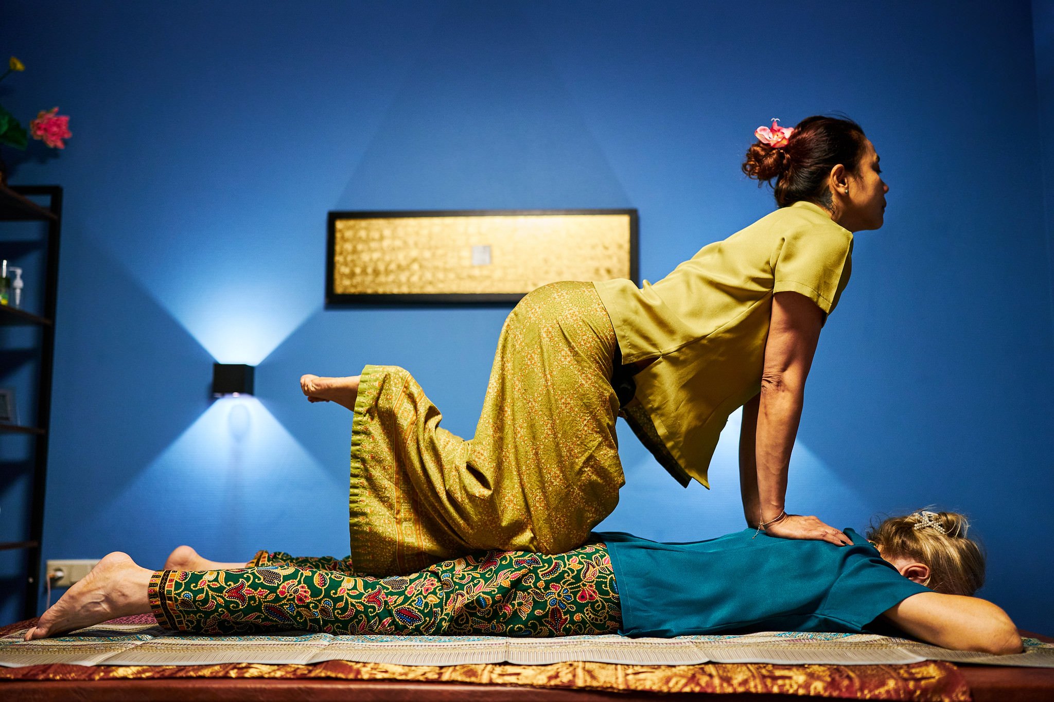 A woman in yellow traditional clothing practicing yoga on a mat with a colorful pattern, with another person lying underneath her, supporting her pose, in a room with blue walls and modern decor.
