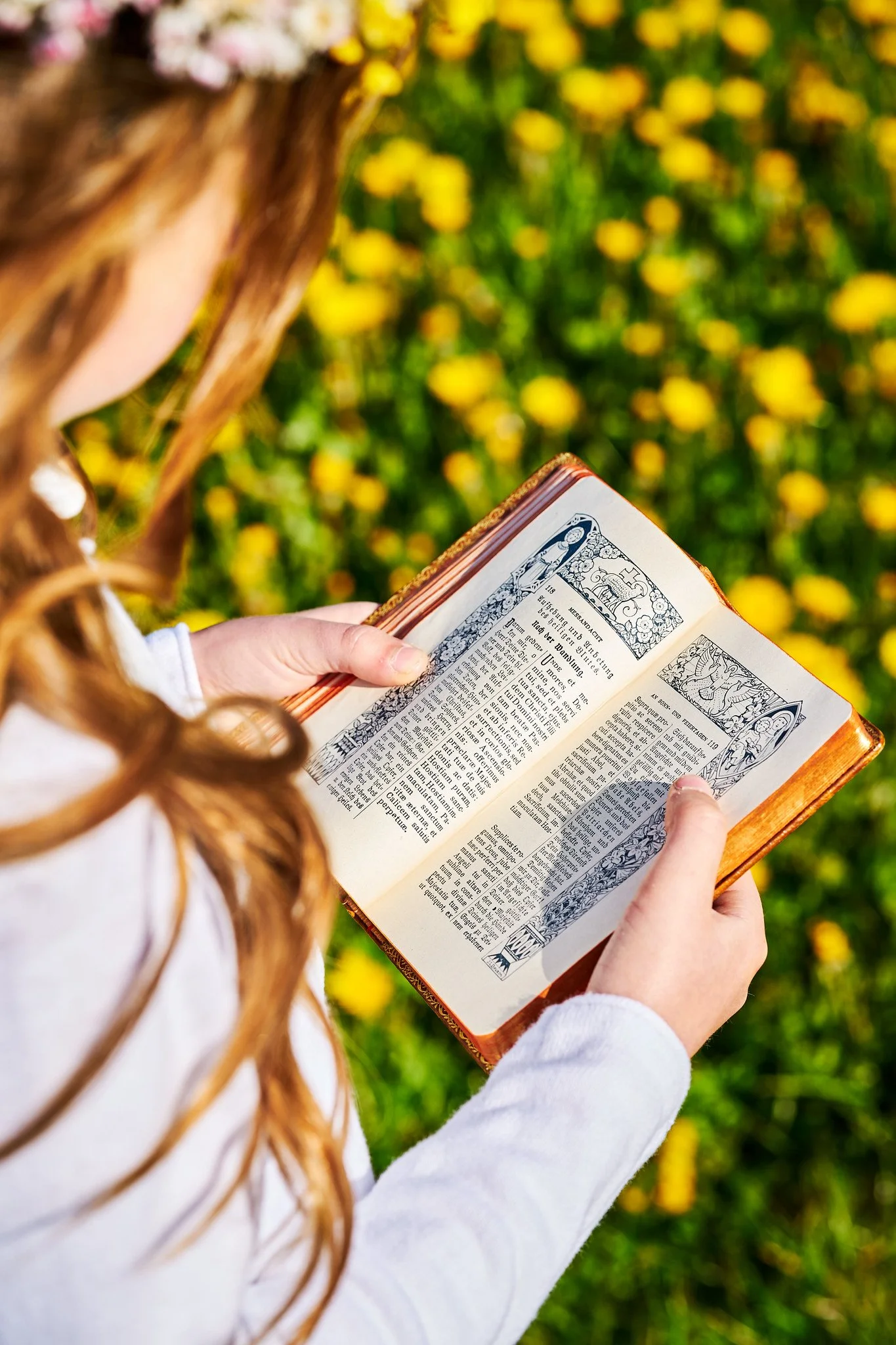 A woman with long red hair holding an open religious book outdoors, with a blurred background of yellow flowers.
