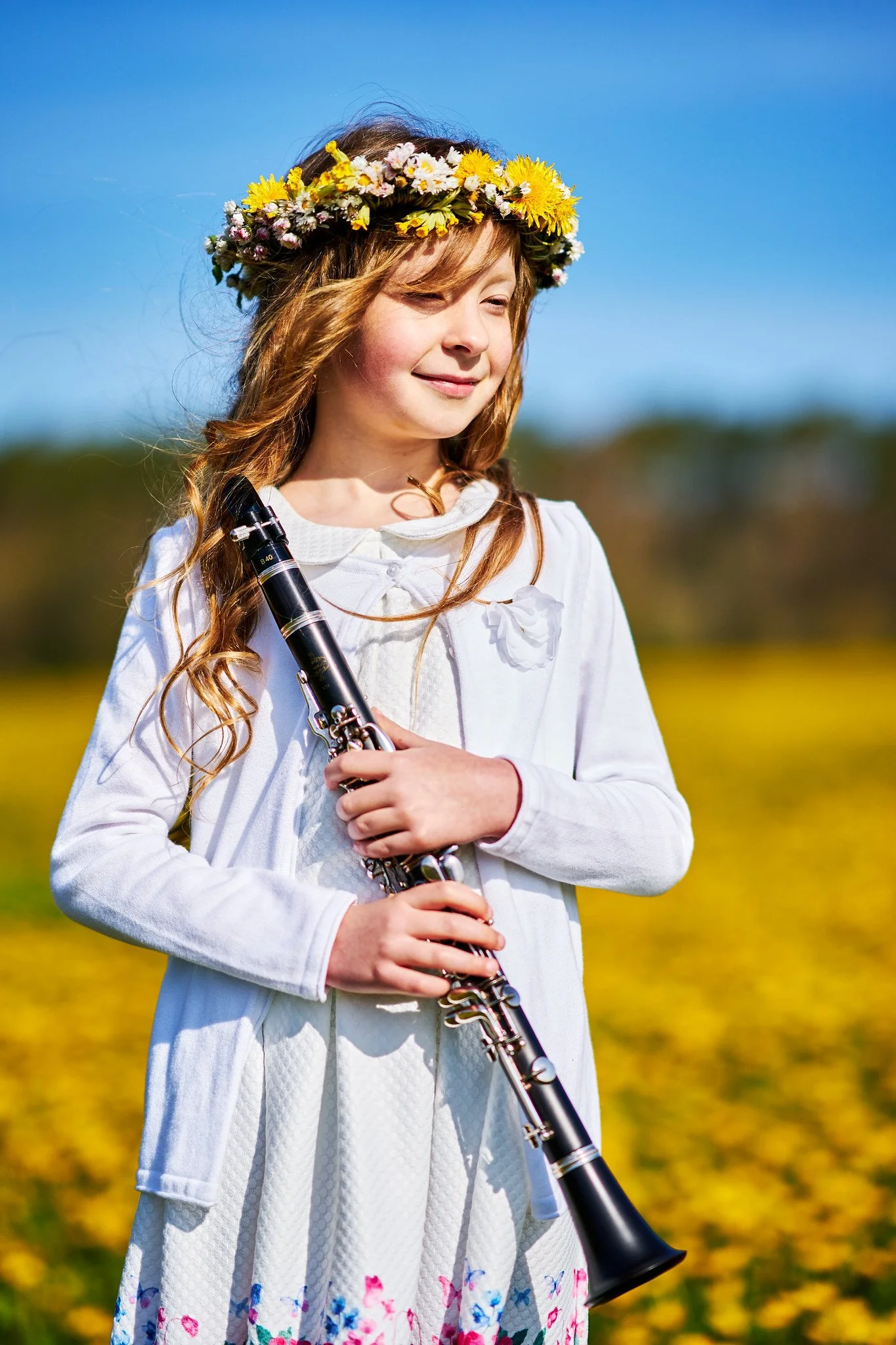 Young girl outdoors wearing a white dress with floral embroidery, a flower crown, and holding a clarinet, with a field and blue sky in the background.