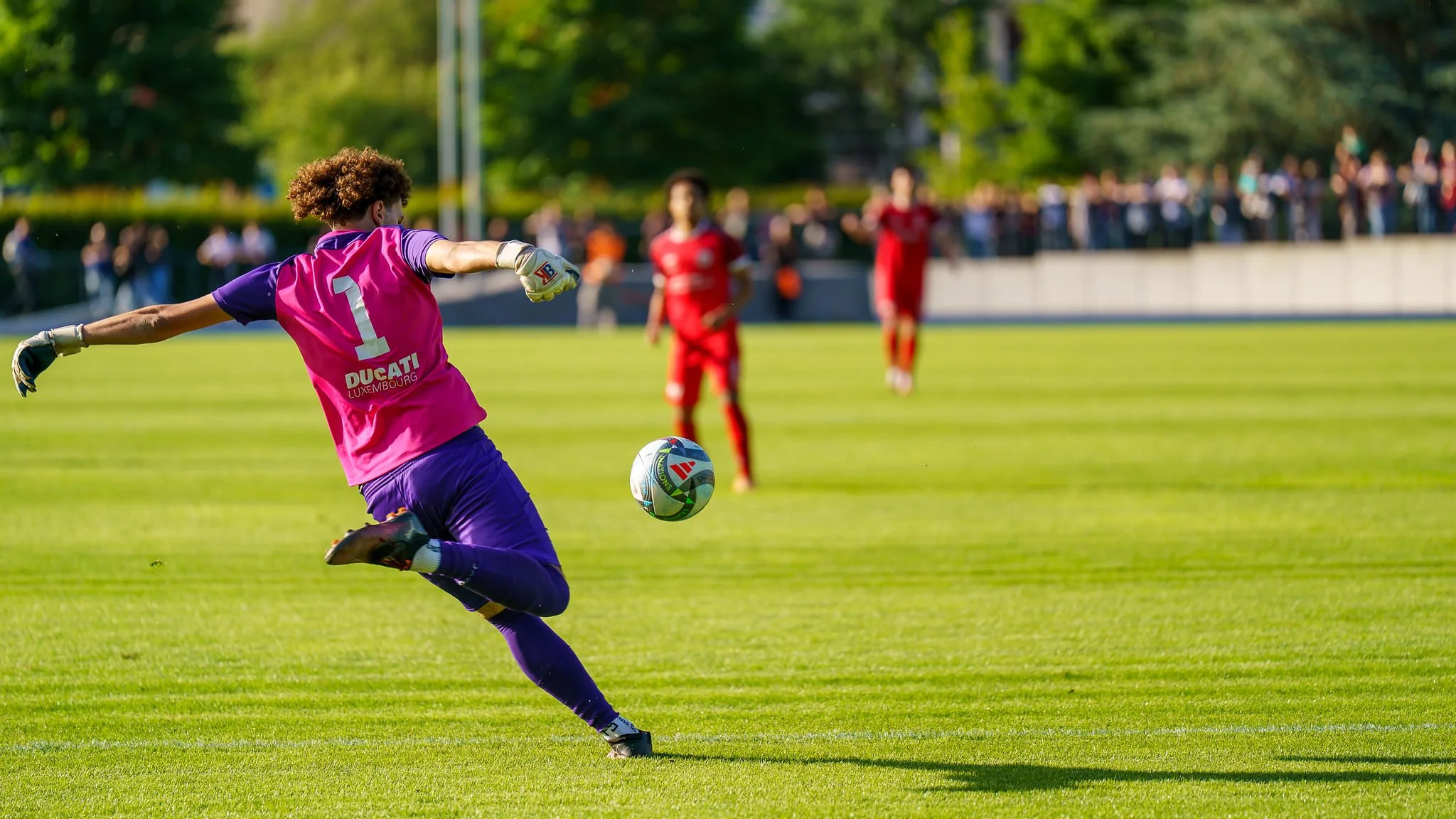 A soccer player wearing a pink and purple uniform kicking a soccer ball on a green field with spectators in the background.