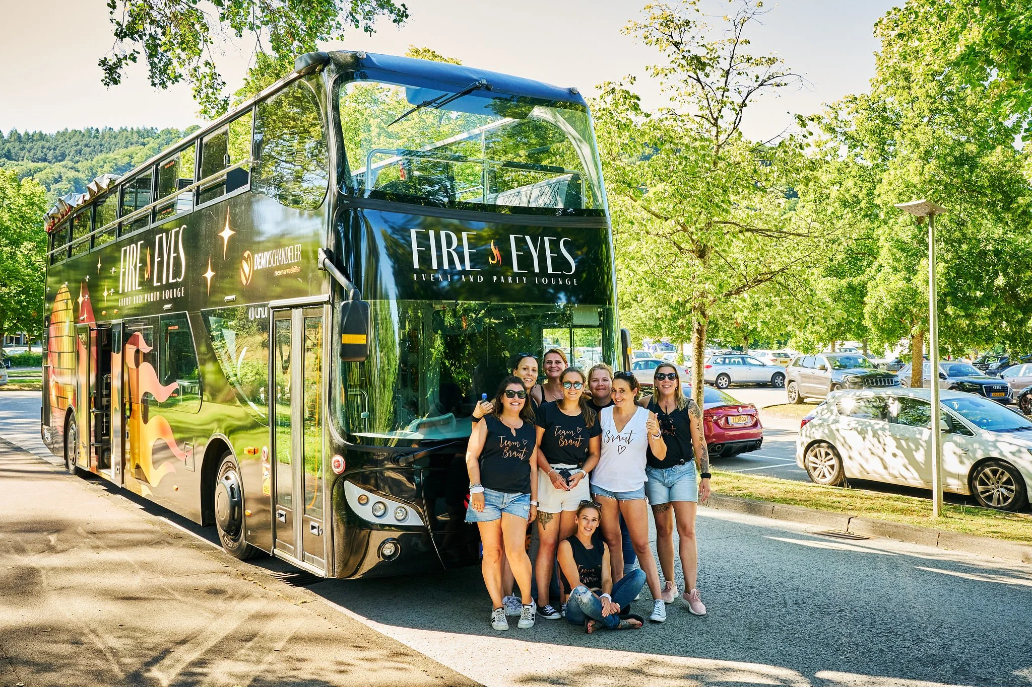 A group of women posing for a photo in front of a double-decker bus labeled 'Fire Eyes Event and Party Lounge,' parked in a lot with trees and other cars around.