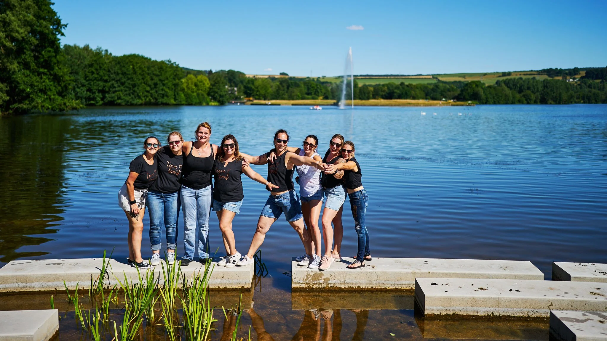 Group of women standing on a dock in front of a lake with scenic water fountains and a lush green landscape under a clear blue sky.